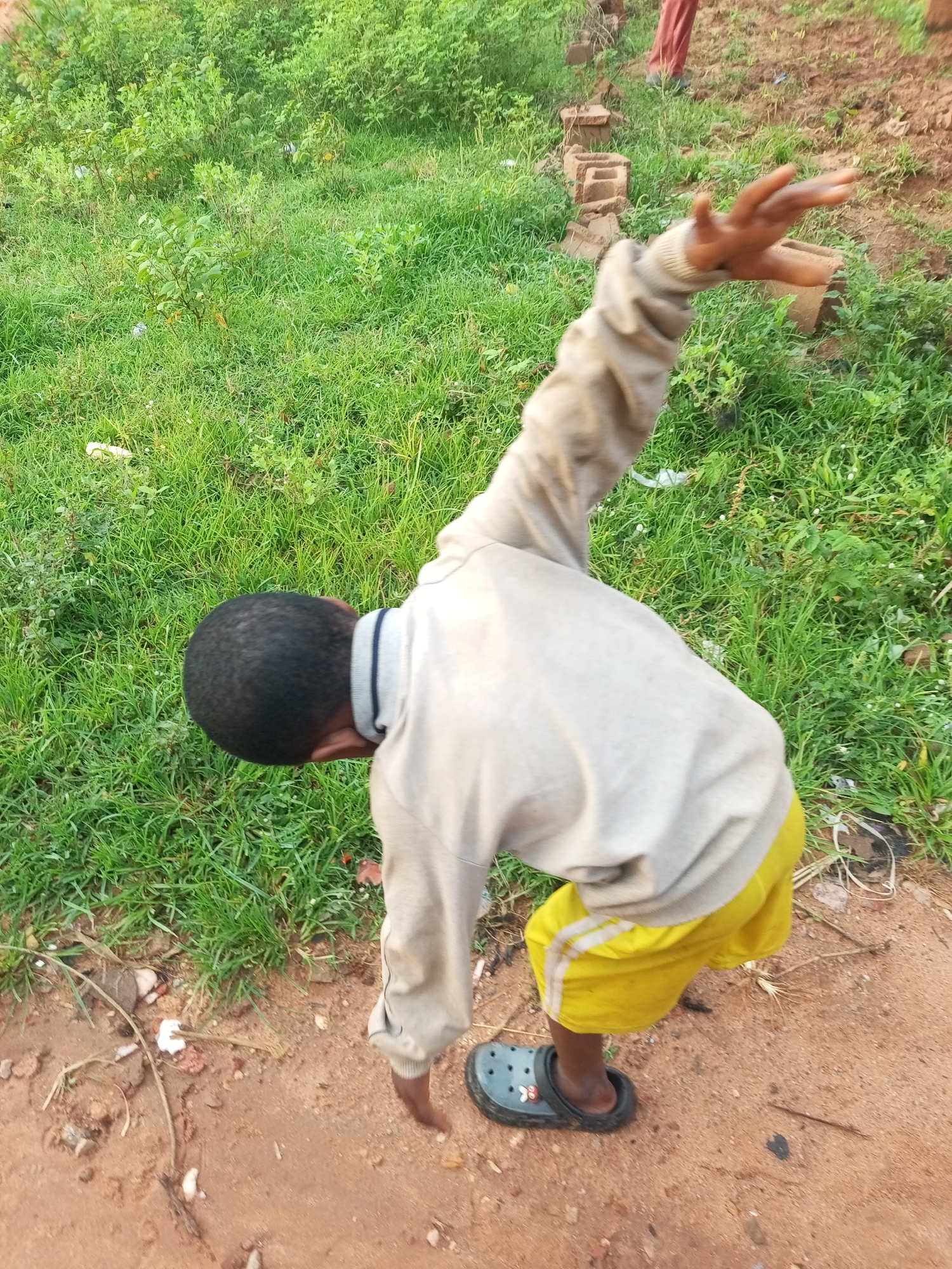 A young boy is captured from a high angle, engaged in an athletic pose outdoors in what appears to be a dusty, unpaved area with lush green grass and some vegetation bordering the path. The boy is wearing a light grey long-sleeved shirt with blue and white trim on the collar and yellow shorts with white stripes. He has black hair and is wearing dark grey clogs. His right arm is extended upwards and to the side, with his hand open. The background features more grass and some scattered bricks. The setting suggests a rural or less developed urban area within Tempure, Nigeria.