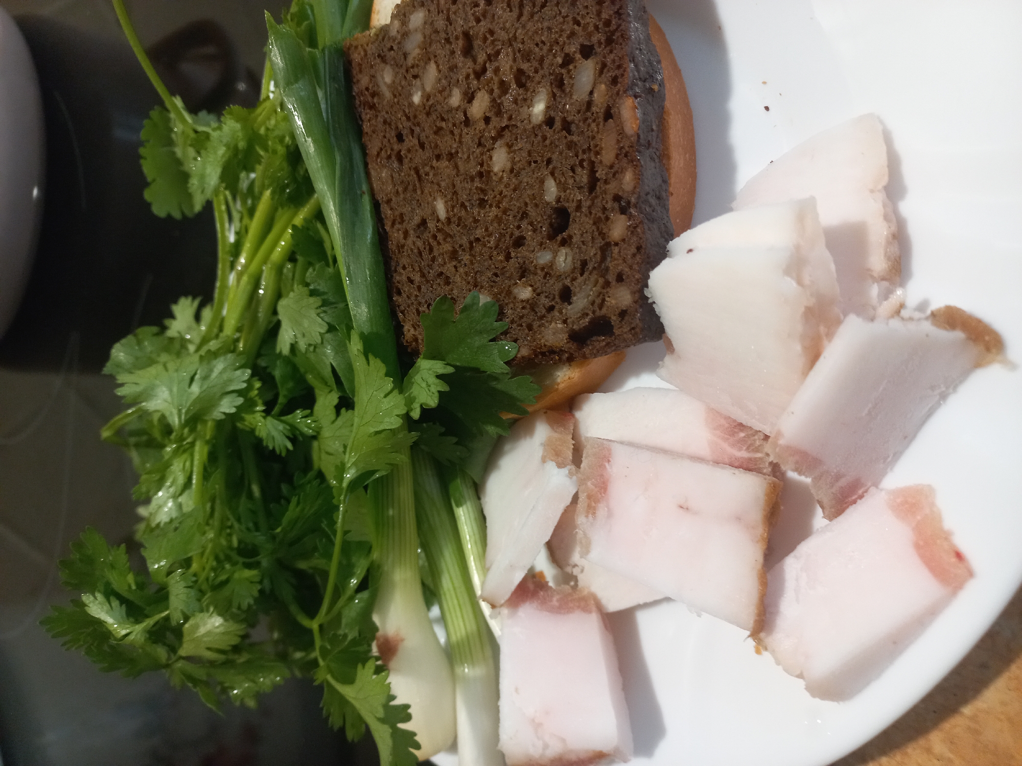 The image depicts a plate of food. There is a piece of dark brown bread with seeds, a bunch of green onions and cilantro, and several slices of what appears to be salted pork fat (salo). The food is arranged on a white plate. The context and items displayed are suggestive of food commonly consumed in Ukraine and Eastern Europe. There is nothing in the image that gives the location as Kyiv.