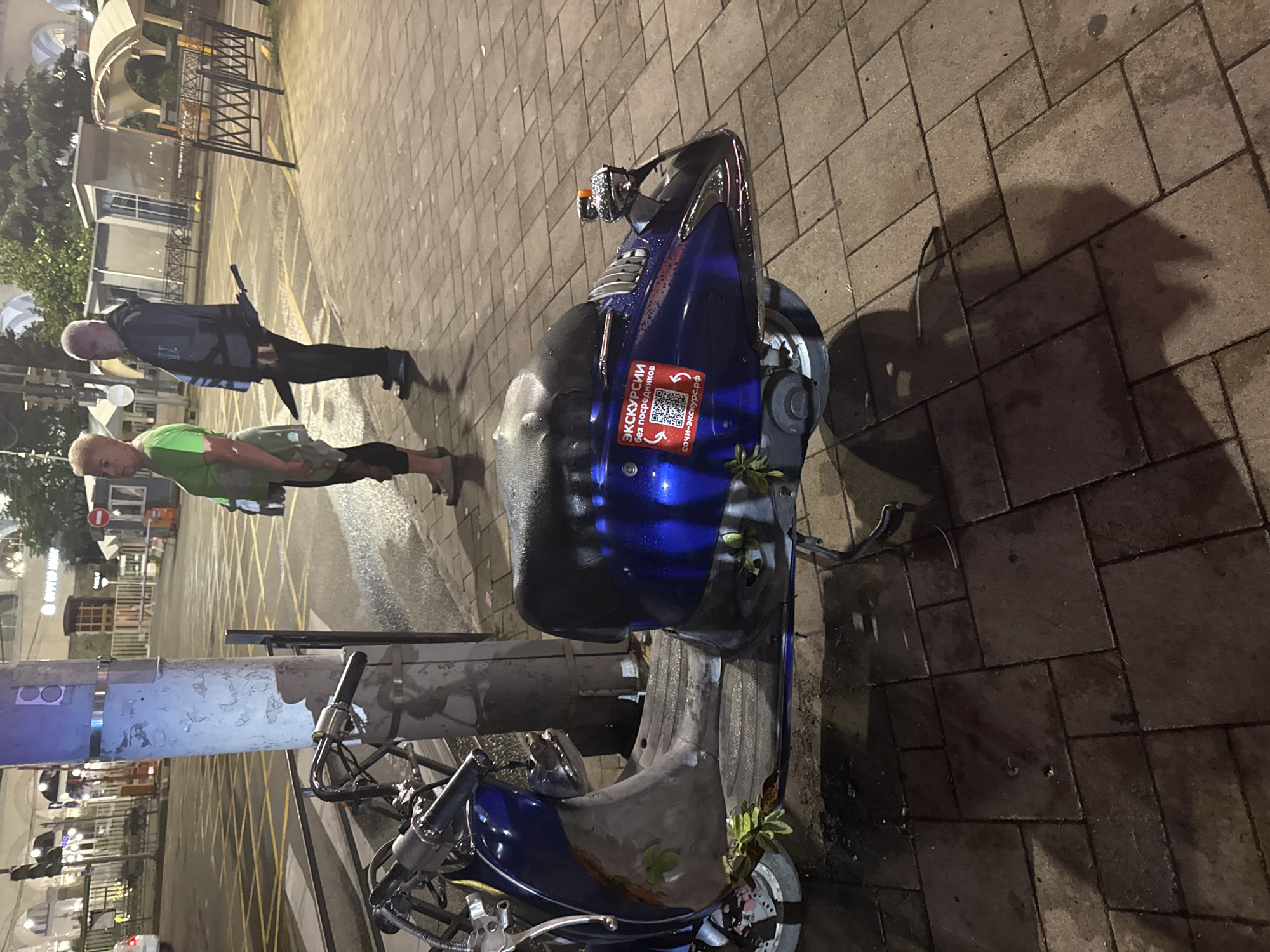 In Sochi, Russia, at night, a blue scooter is parked on a paved sidewalk adjacent to a street with yellow road markings. Two people, a woman wearing a green shirt and dark pants, and a man in a dark jacket and pants, stand on the sidewalk. The man is holding a long, dark object, possibly an umbrella or a blade, tucked behind his leg. The scooter has a sticker with Russian text and a QR code on its side. The environment is lit by streetlights, casting shadows on the ground.