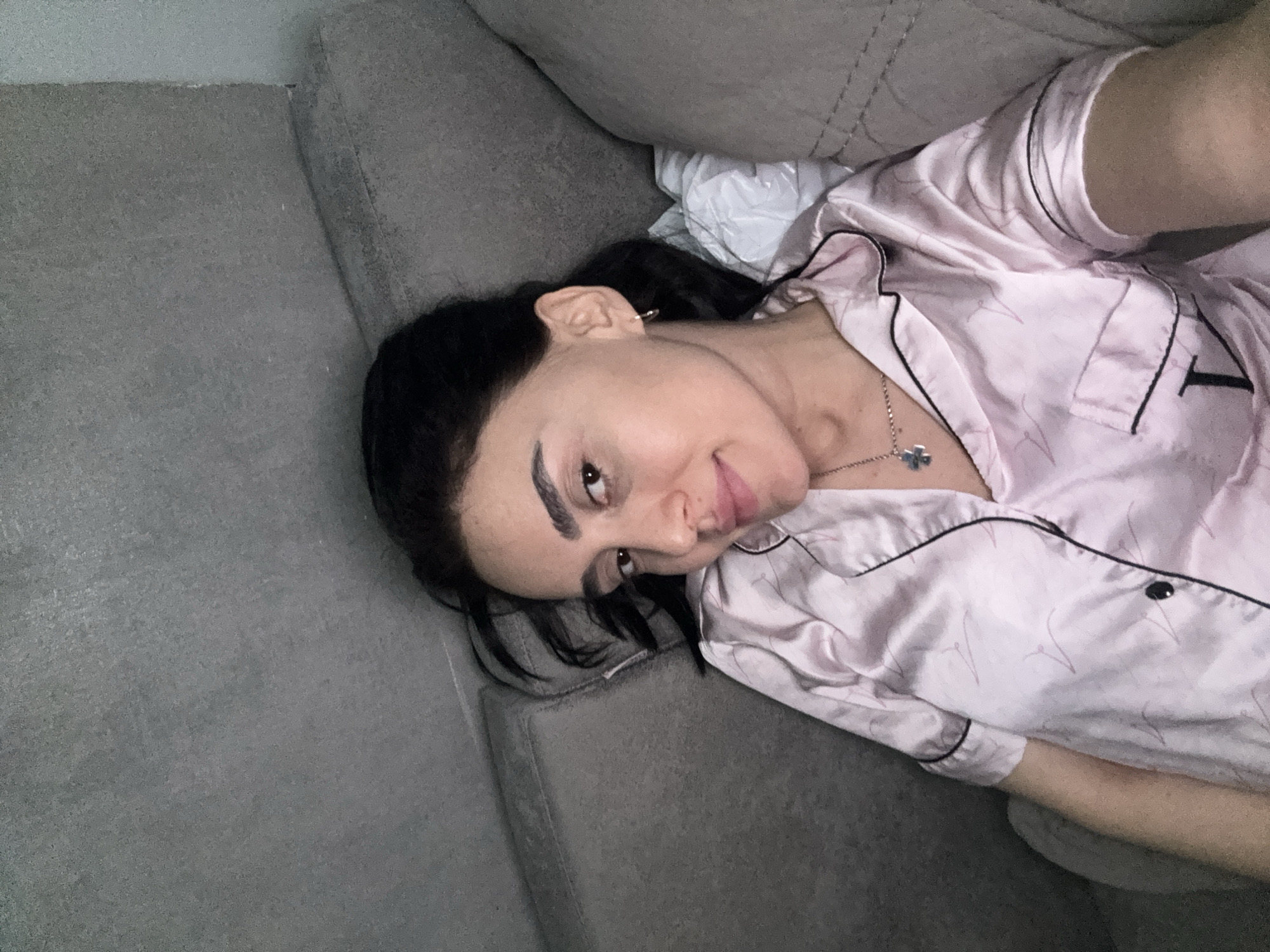 A woman is lying on a grey sofa. She has dark hair and is wearing a pink pajama set with black piping. She has a silver necklace with a four-leaf clover pendant. The location is not discernable.