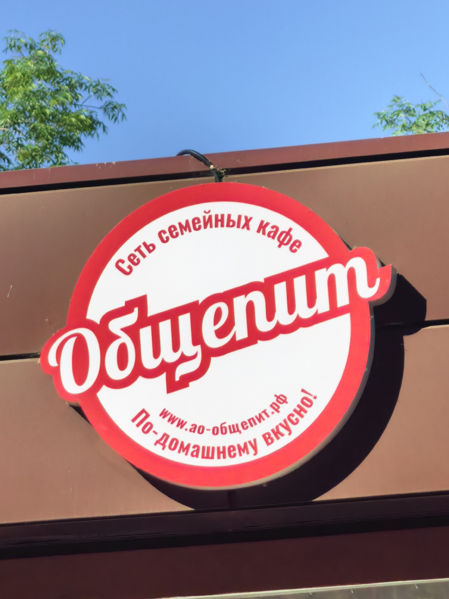 The image shows a sign for a cafe in Sterlitamak, Russia. The sign is circular with a red border and white background. It features the name "Общепит" in large, stylized red letters. Above the name, in smaller white letters, is the phrase "Сеть семейных кафе" (Network of Family Cafes). Below the name, in red letters, is "По-домашнему вкусно!" (Homemade tasty!) along with "www.ao-общепит.рф". The sign is mounted on a brown building facade, with green foliage visible in the upper left and right corners of the frame. The sky is clear blue.