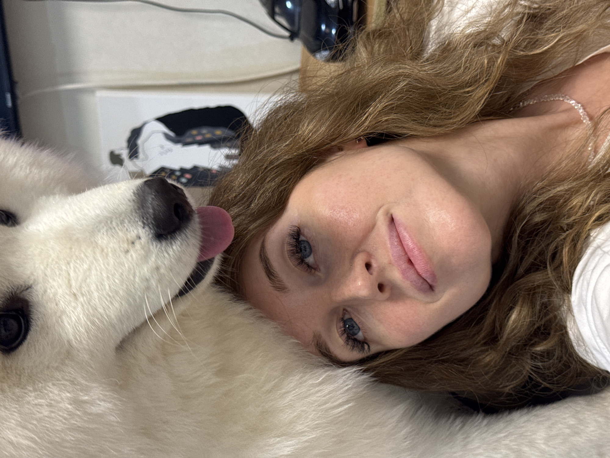 The media file is a close-up, low-angle photograph taken indoors, possibly in a bedroom or living room, featuring a young woman and a white dog. The woman is lying on her back, with her head tilted and resting on a white furry surface, likely a rug or blanket. Her eyes, which are blue, are looking towards the camera. She has long, wavy brown hair spread around her face and shoulders. Her makeup includes noticeable eyelashes and lip gloss. A thin, sparkling necklace is visible around her neck. The dog, a white fluffy breed, is positioned on the left side of the frame, its snout close to the woman's face. The dog's tongue is slightly out, and its nose is black. In the background, out of focus, there appears to be some electronic equipment, including a black device with lights and a white surface with some patterned detail. The lighting suggests it is daytime, but there are no strong indicators of the weather or specific time of day. The overall mood of the image is relaxed and affectionate, with the close proximity of the woman and her pet suggesting a moment of comfort and companionship. There is no visible text in the image. The context provided indicates the location is Chaikovsky, Russia.
