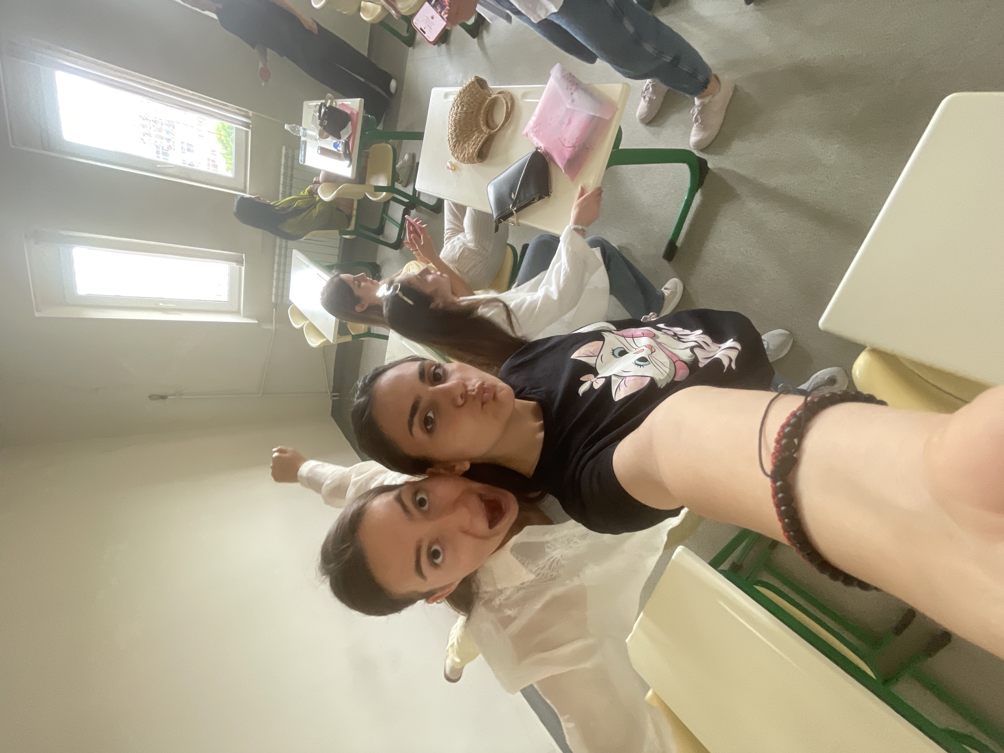 The image depicts a casual scene among several young women in what appears to be a classroom or training room in Burgas, Bulgaria. In the foreground, two individuals are taking a selfie. One woman, wearing a black t-shirt adorned with a white cartoon cat design (Marie from Aristocats), pouts playfully towards the camera. Beside her, another woman in a white long-sleeved shirt displays an exaggerated open-mouthed expression. A red and black beaded bracelet is visible on the arm of the woman in the black t-shirt.
In the background, other individuals are seated at light beige desks with green metal frames. Some are casually engaged with their mobile phones. Various personal items are visible on the desks, including a straw bag, a black clutch bag, a pink translucent bag, and a water bottle. The room is well-lit by bright natural light streaming through two rectangular windows on the left side of the frame, indicating it is daytime. The walls are a light neutral color, and the floor is a light greyish-beige. The overall atmosphere suggests a relaxed, informal gathering or a break during a session.