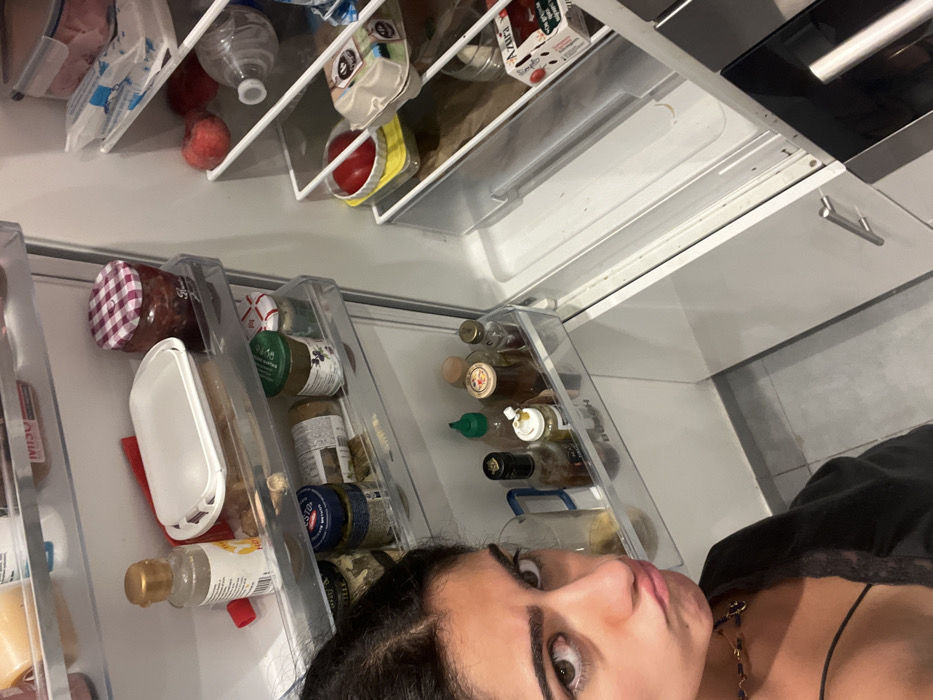 A refrigerator's door shelves display various items: condiments like small jars of honey, pickles, and sauces are visible. The main compartment contains apples, bottled water, and packages of what appears to be lunch meat and tomatoes. A woman's face is prominently in the foreground, partially obscuring the lower shelves. She wears a dark top, and her gaze is directed towards the viewer. The refrigerator itself has white interior panels.