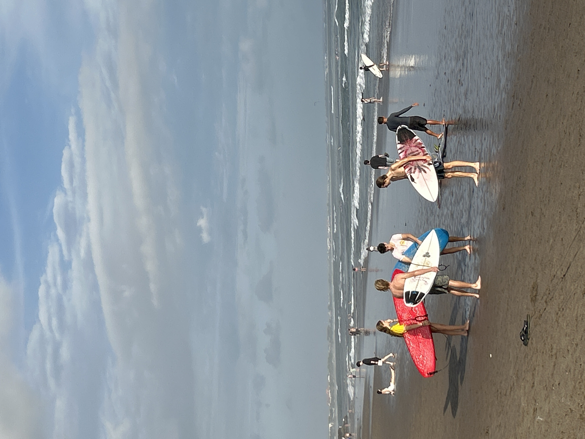 An overcast day at the beach in North Kuta, Indonesia, features a group of people interacting with surfboards on the shoreline. The foreground is dominated by the wet sand of the beach, with a pair of black flip-flops lying abandoned near the center.
To the left, a young woman in a yellow top stands next to a bright red surfboard, while a blonde-haired young man holds a white surfboard with blue accents. To their right, another young man in a white t-shirt stands beside them.
Further down the beach, towards the water's edge, a young man with a patterned surfboard walks towards the ocean. Behind him, another individual in dark attire is partially obscured by the surf. In the middle ground, several other figures are visible, some standing in the shallow water, others walking along the wet sand.
The ocean extends into the background, with moderate waves breaking and receding. Further out, numerous small figures can be seen in the water, likely surfers. The horizon is marked by a hazy line where the sea meets the sky. The sky itself is a canvas of diffused light, with a mix of grey clouds and patches of pale blue. The overall atmosphere is one of relaxed, casual beach activity.