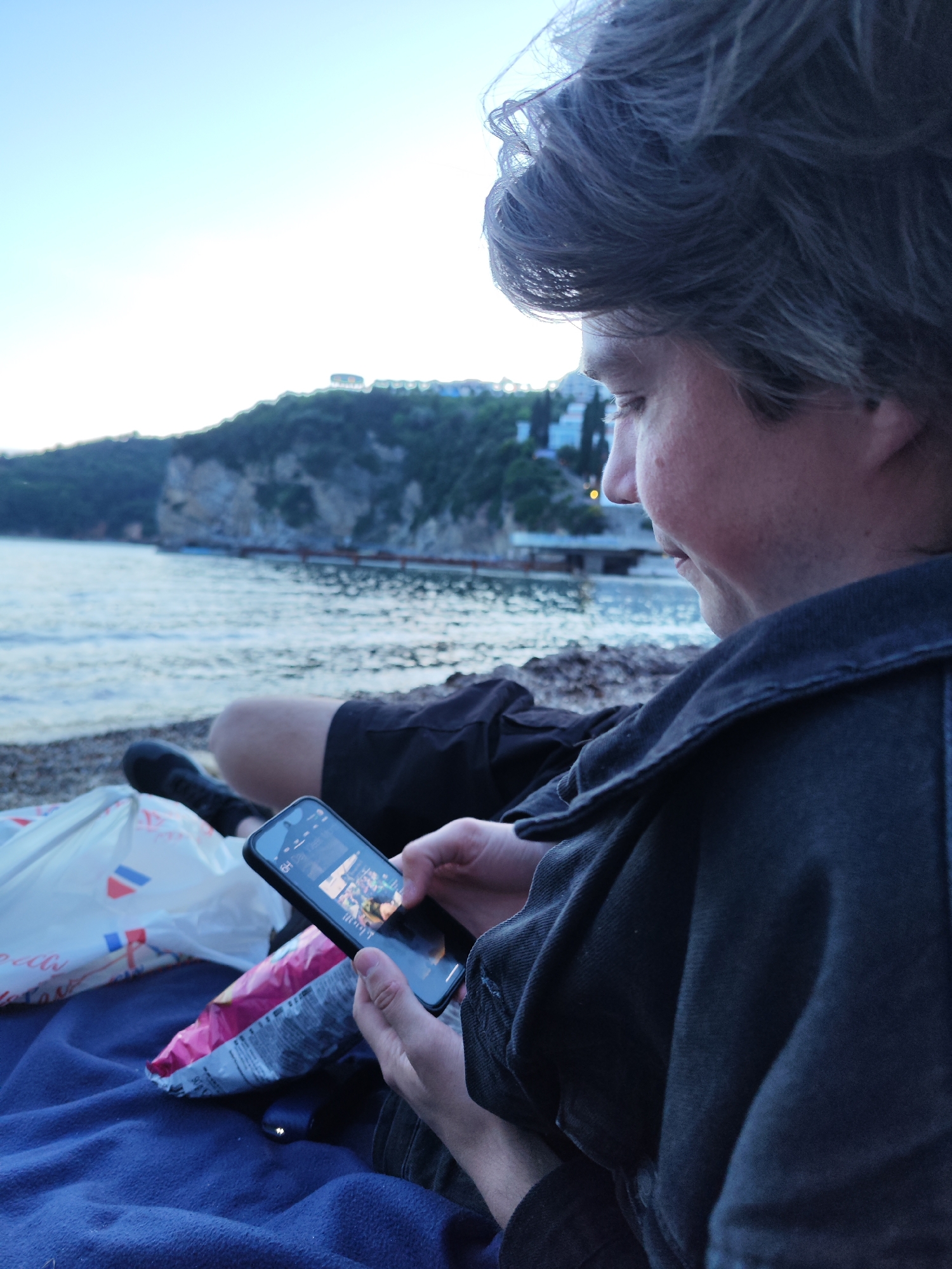 A man is sitting on a dark blue blanket on a beach, looking at his smartphone. He has short gray hair and is wearing a black denim jacket. Next to him is a white plastic bag and a bag of chips. In the background, there is a body of water with a rocky land mass with trees on top. The location is Budva, Montenegro.