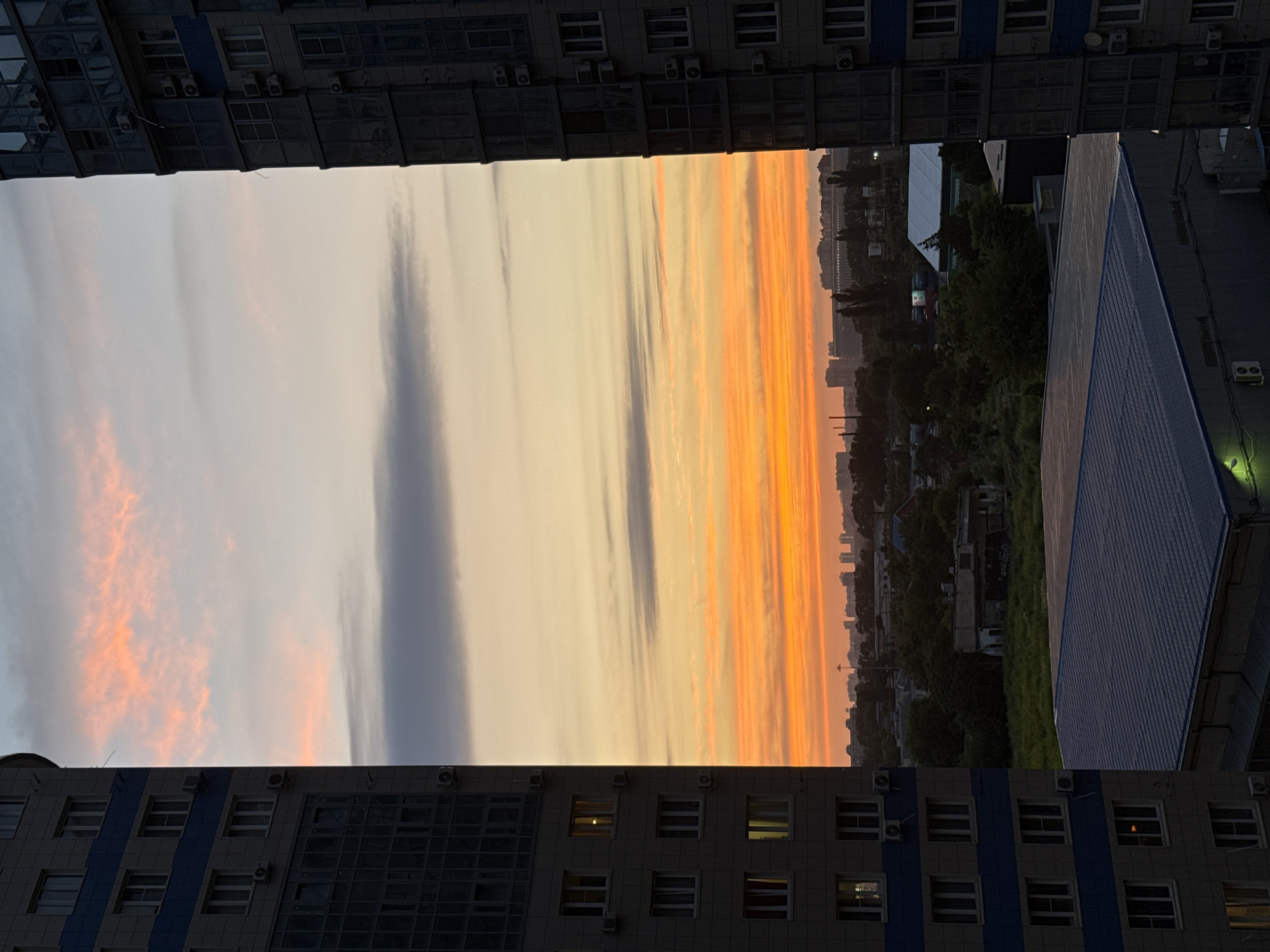 The image captures a sunset over the city of Krasnodar, Russia. The sky is a vibrant spectacle of orange, yellow, and pink hues, with streaks of grey clouds interspersed. Two tall apartment buildings flank the scene, their dark facades contrasting with the bright sky. One building is on the left, its windows showing lights within, suggesting occupancy. The other building is on the right, also dark and imposing. Between the buildings, a cityscape unfolds with various structures, including lower-rise buildings, trees, and hints of industrial elements. The foreground features a portion of a flat-roofed building, its metallic surface reflecting the dim light. The overall impression is an urban panorama at dusk, a common sight in a Russian city.