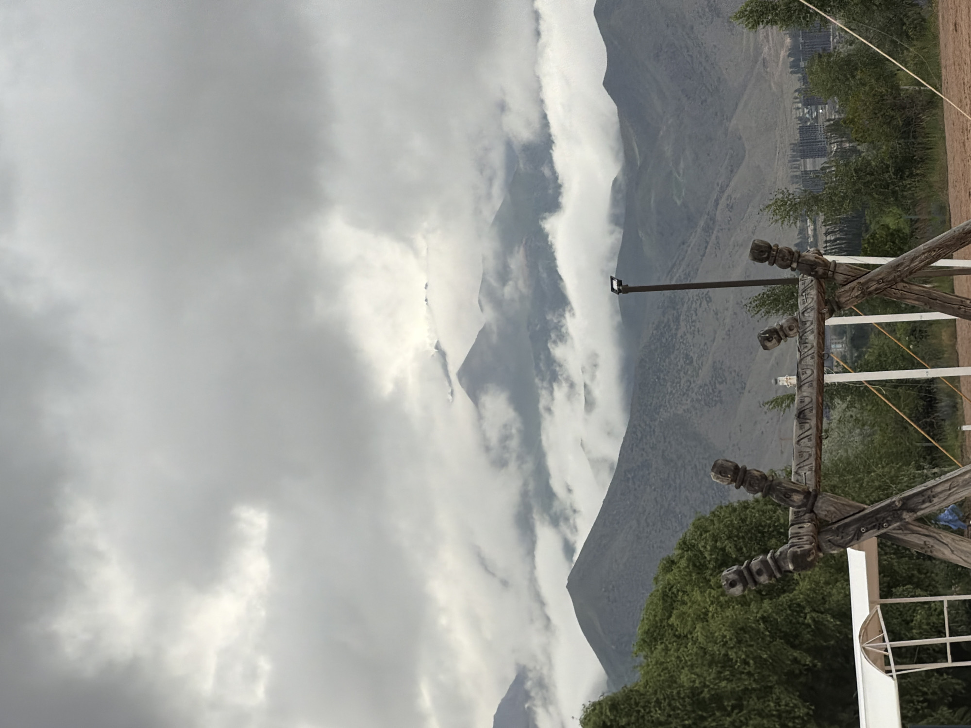 The image depicts a cloudy day with a mountain range in the background. In the foreground, a wooden swing set with carved details is visible. A white canopy structure is partially visible to the left of the swing set. Trees and vegetation surround the area. In the distance, a city with several tall buildings can be seen nestled among the hills. The scene appears to be an outdoor recreational area in or near Çolpon-Ata, Kyrgyzstan, given the mountain landscape. There are no people visible in the image. The lighting suggests it might be early morning or late afternoon due to the overcast conditions.