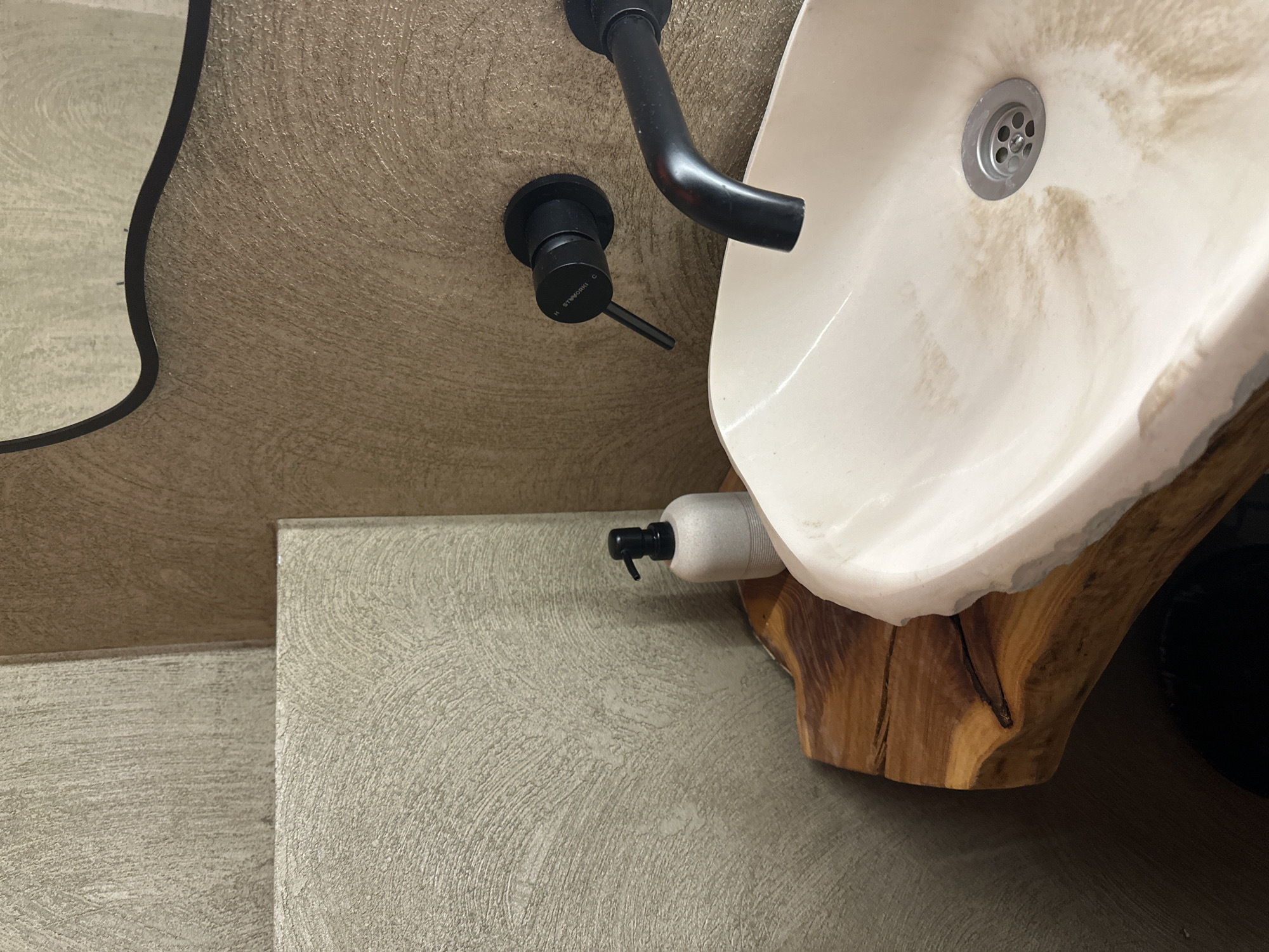 This image depicts a detailed view of a contemporary bathroom sink area, located in Усады, Russia. A prominent white ceramic vessel sink, displaying significant light brown and beige discoloration or staining within its basin, is the central object. A circular silver drain grate is visible at the bottom of the sink. The sink is supported by a live-edge wooden slab countertop, characterized by its natural brown tones, distinct wood grain, and visible cracks. Above the sink, a matte black wall-mounted faucet assembly consists of a curved spout and a single lever mixer handle, on which the text "H STIEWERKI C" is discernible. To the left of the sink, resting on the wooden countertop, is a light grey or beige cylindrical soap dispenser with a black pump. The surrounding walls are textured with a swirled or brushed pattern. The wall behind the faucet is light brown, while adjacent wall sections to the left feature a similar textured pattern in a lighter, greyish-beige hue, appearing as distinct panels. A segment of a mirror with a dark, wavy frame is visible in the upper right portion of the image, reflecting a portion of the textured wall.