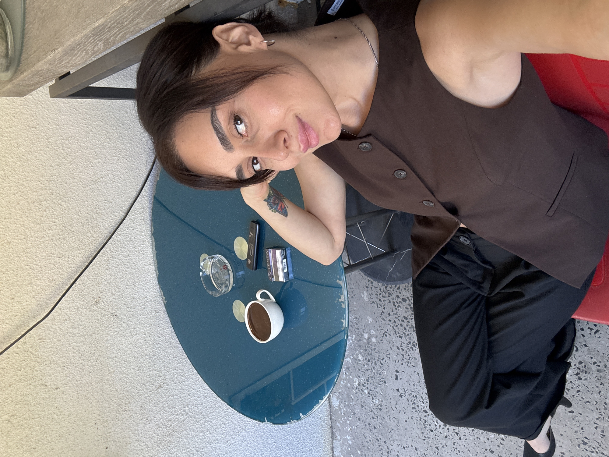 A young woman with dark hair is sitting at a blue table outdoors. She is wearing a brown vest over a black shirt and black pants. On the table, there is a cup of coffee, an ashtray with a cigarette butt, a pack of cigarettes, and a lighter. A butterfly tattoo is visible on her left arm. The background consists of a white textured wall and a gray speckled ground. The scene appears to be a cafe or outdoor seating area.