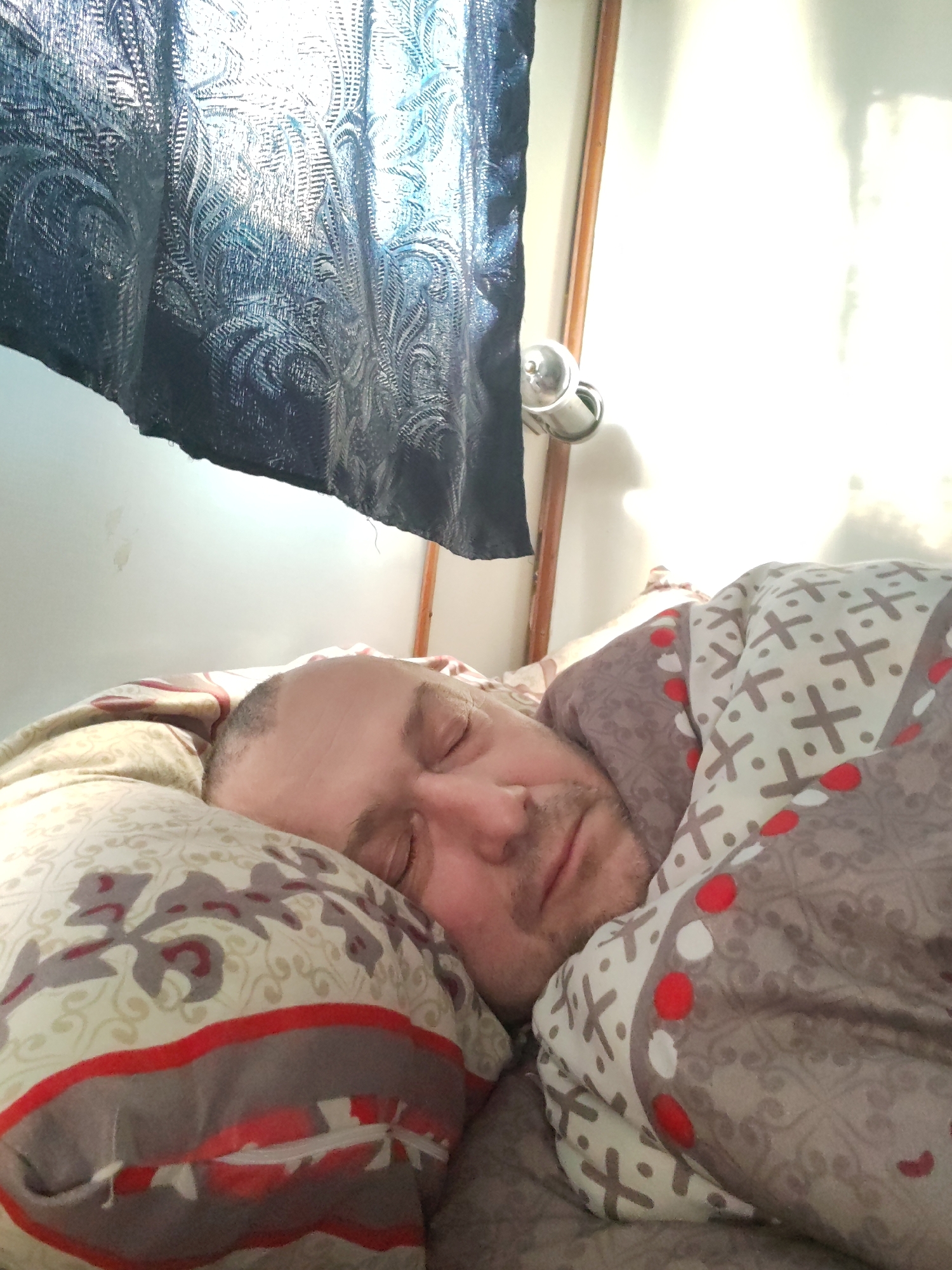 This close-up indoor photograph depicts a man sleeping peacefully in bed. The man, who appears to be middle-aged with short hair and some facial stubble, is lying on his side with his eyes closed. He is partially covered by a duvet featuring a pattern of gray crosses and red and white dots on a cream background. His head rests on a pillow adorned with a floral or ornamental design in shades of red, gray, and cream.
Bright natural light streams into the room from the left side of the frame, strongly backlighting a dark, possibly navy or black, patterned curtain that hangs in the upper left corner. The intensity of the light creates some overexposed areas on the light-colored wall to the right. A silver door handle or wall-mounted fixture is visible on the right, attached to a wooden frame.
The scene suggests a bedroom setting, likely during the daytime, possibly morning, given the strong, direct sunlight illuminating the space and the individual's state of sleep. The man's relaxed posture and closed eyes convey a sense of calm and rest. There is no visible text in the image.