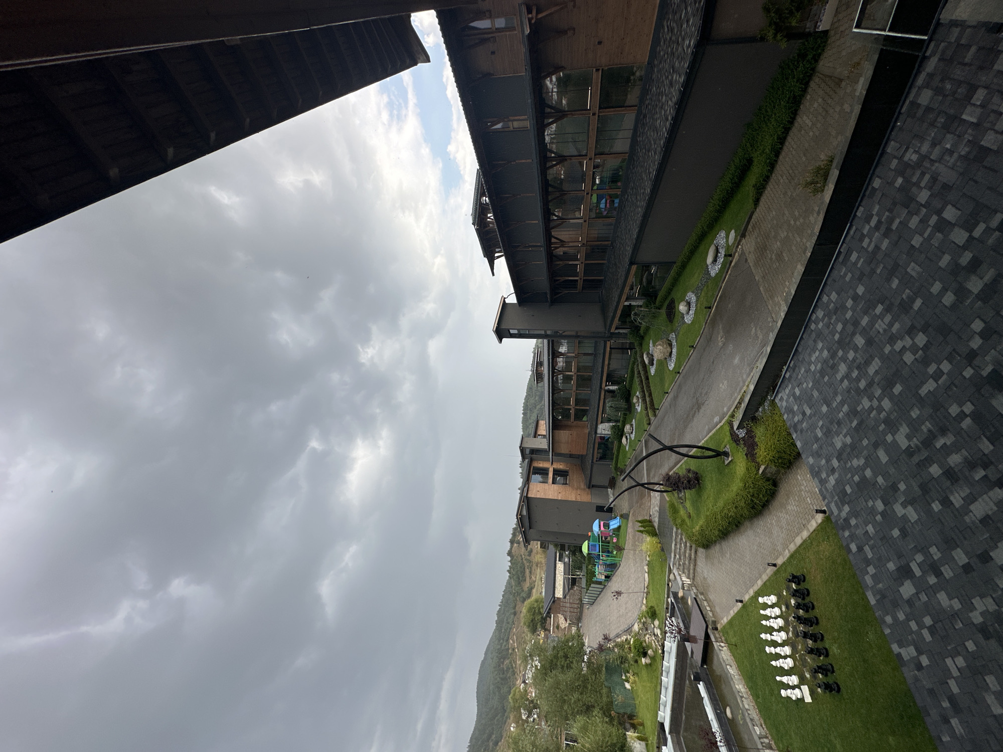 The image depicts an outdoor scene in the city of Bania, Bulgaria, characterized by a cloudy sky and a modern hotel or resort complex. The architecture features dark gray and brown wooden exteriors with large glass windows and balconies. In the foreground, a rooftop with dark gray shingles is visible. Below, a paved walkway leads through a well-maintained garden area with manicured lawns, decorative rocks, and shrubs. A large chess set with black and white pieces is arranged on the lawn. Further into the complex, a children's playground with colorful slides and climbing structures is situated. In the background, rolling hills covered in trees suggest a mountainous or hilly terrain. The overall atmosphere is overcast and somewhat muted due to the weather conditions.