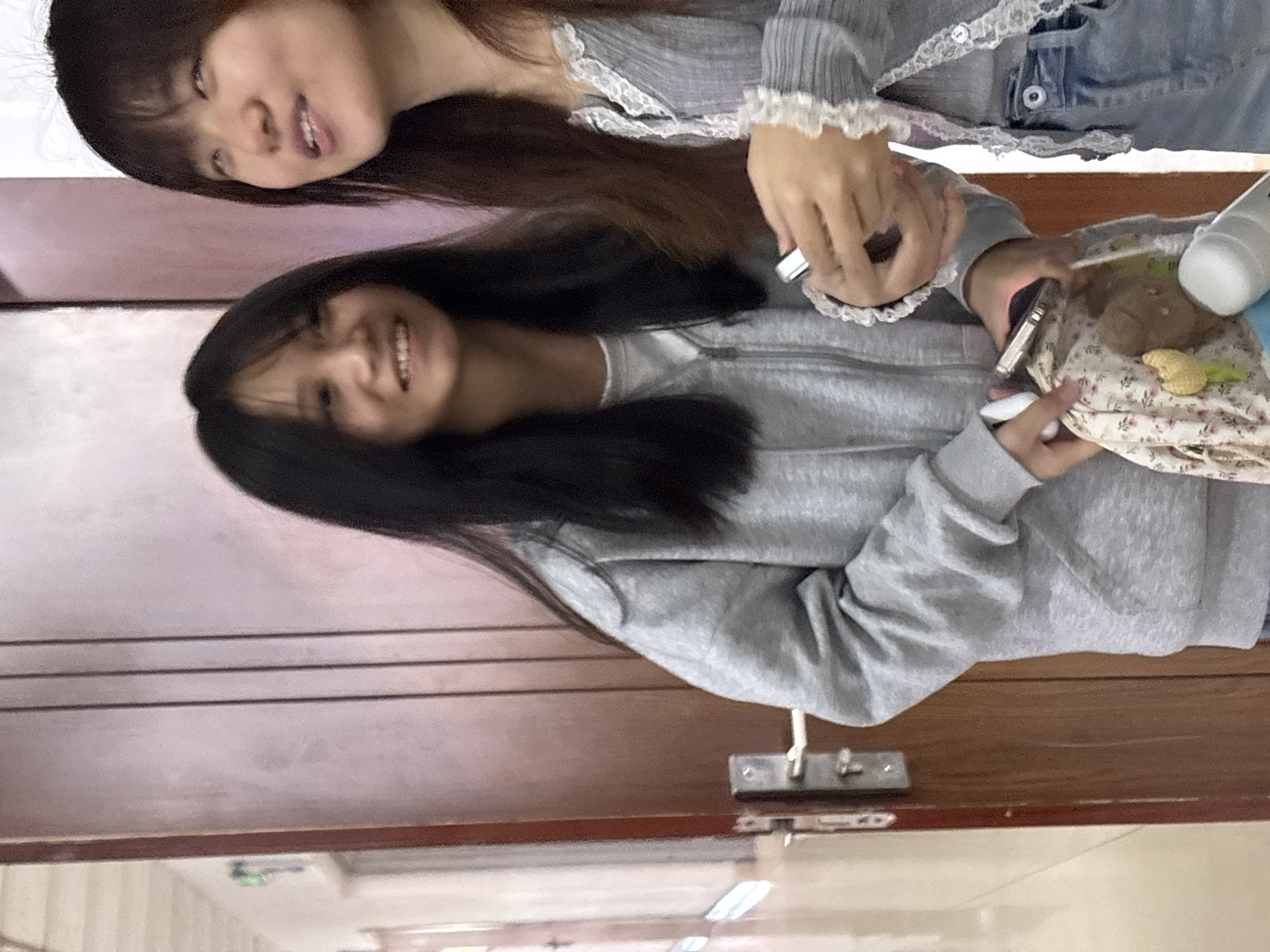 Two young women are posing for a photo indoors, likely in a home or dorm room. The woman in the foreground has long black hair and is wearing a gray sweatshirt. She is smiling and looking at the camera while holding a collection of items, possibly related to self-care or beauty products, in her hands. Beside her is another woman with long brown hair and a gray cardigan with lace details, smiling into the camera too. A brown wooden door and some flooring are visible in the background. The location cannot be determined from the image.