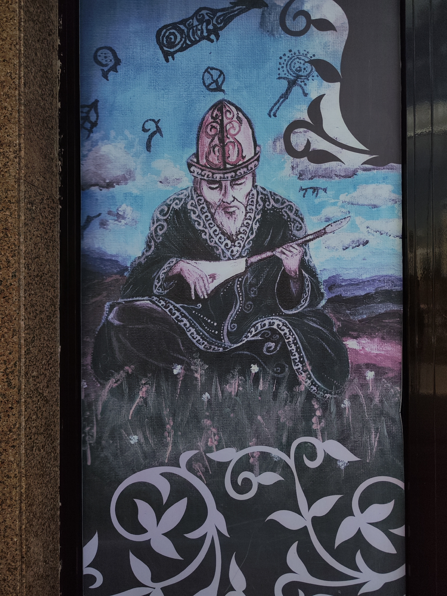 This is a vertical, outdoor photograph of a large mural or poster depicting a seated man in traditional attire playing a stringed instrument. The man has a beard and wears an ornate, segmented hat. He is dressed in a dark, embroidered robe. The background of the mural is a painterly depiction of a sky with clouds and distant, rolling hills in shades of purple and pink. Scattered across the sky are several abstract, black symbols and figures, some resembling ancient script or stylized animals. The foreground of the mural shows a grassy field with scattered white flowers and a decorative, white, stylized vine pattern at the bottom. The mural appears to be attached to a building with a textured, beige wall on the left and a dark, reflective surface on the right, possibly a doorway or window frame. The image was captured in Astana, Kazakhstan, likely in an urban setting given the architectural elements.