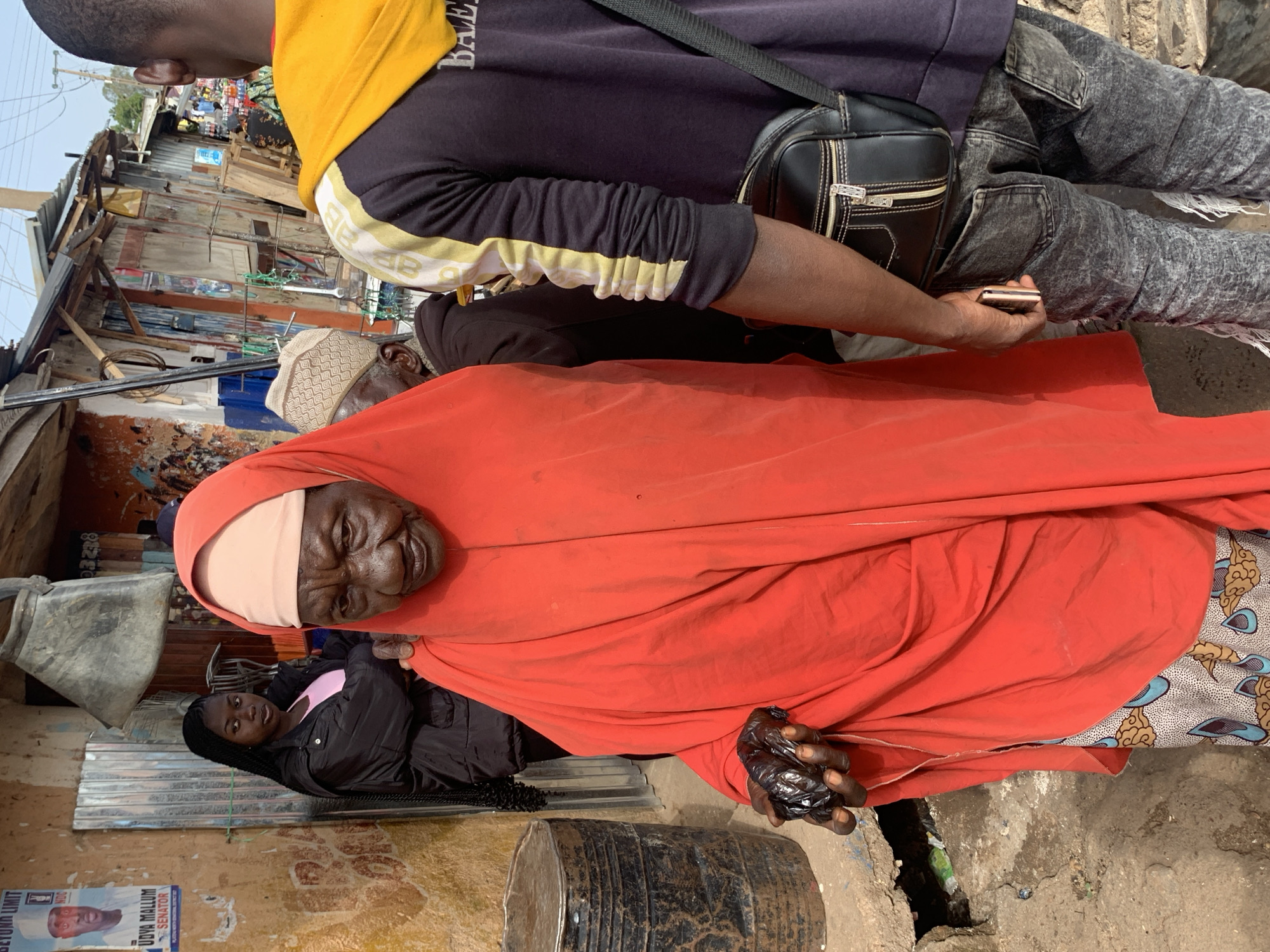 A Nigerian woman, elderly, wears an orange head covering and dress. She stands in a street, her dark hands holding a black plastic bag. A young woman with braided hair, wearing a black jacket over a pink shirt, stands behind and to her left. A man, partially visible from the back, wears a dark hoodie with yellow and white accents and a black satchel. He holds a mobile phone. The street is lined with structures and a barrel. The scene appears to be in Gura Top, Nigeria.