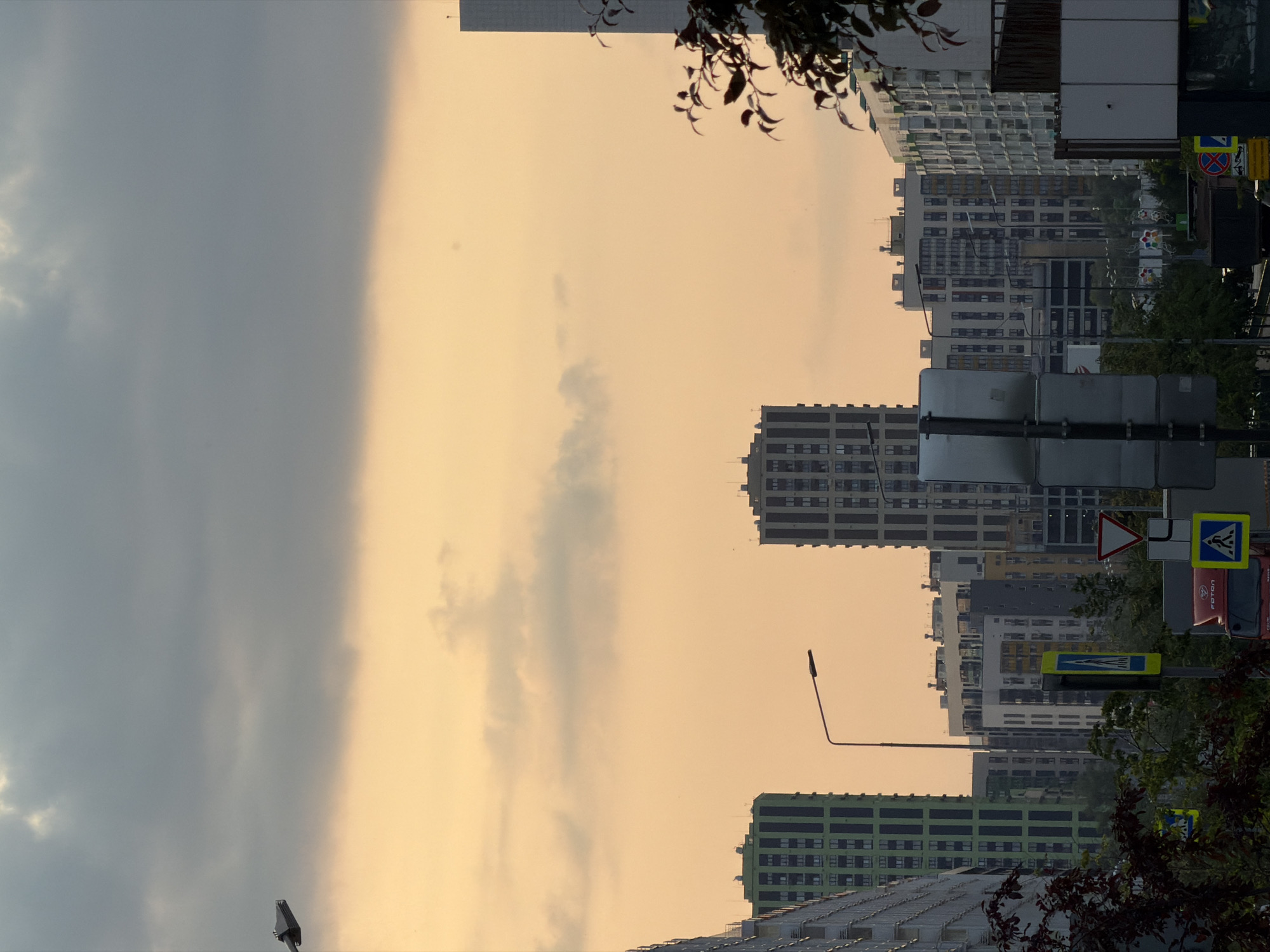 A wide shot shows a city skyline with modern apartment buildings against a cloudy sky with a soft orange glow. The buildings are tall and rectangular, with many windows. Some buildings are green, while others are white or grey. A street lamp is visible in the foreground, and several road signs are on poles. Trees and foliage are also present, adding a touch of nature to the urban landscape. The overall impression is one of a contemporary city at dusk.