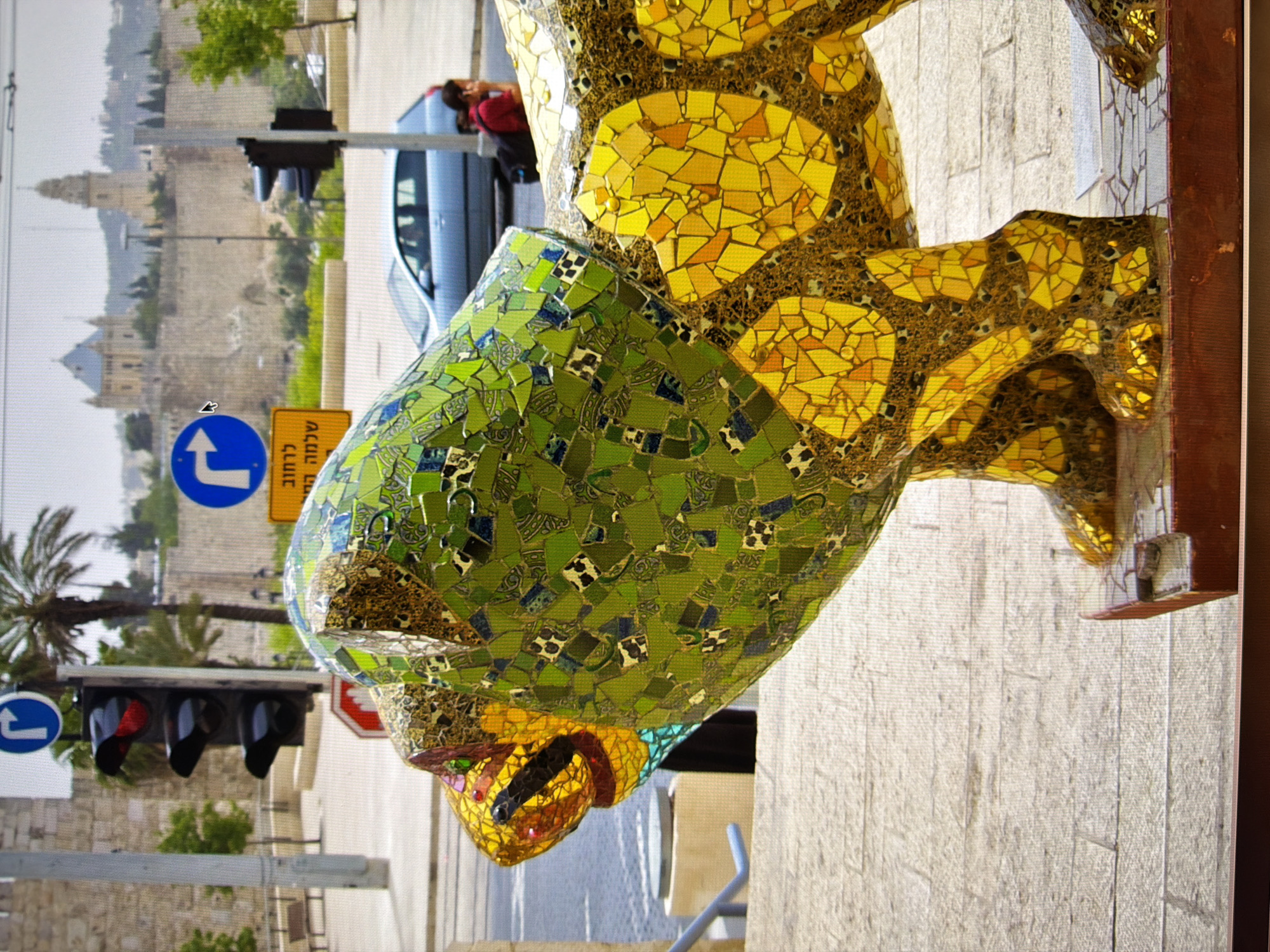 The image shows a mosaic sculpture of a lion's head and upper body. The sculpture is made of many small, colorful tiles. The lion has a green tiled mane with black and white patterned tiles interspersed. Its face and body are tiled with gold and yellow pieces, with red and blue accents around the eyes and mouth. The sculpture is positioned on a stone sidewalk, with a brown wooden base visible at the bottom. In the background, a traffic light with a red light illuminated is visible, as well as street signs and a distant view of buildings with towers and walls, suggesting an urban setting. A blue car is parked on the street behind the sculpture. A person wearing a red top and black backpack is partially visible in the background, to the right of the car. The overall scene depicts an outdoor public space with street elements and urban architecture. The request to include the location in Kyiv, Ukraine, cannot be fulfilled as the image does not provide any visual cues to confirm this specific location.