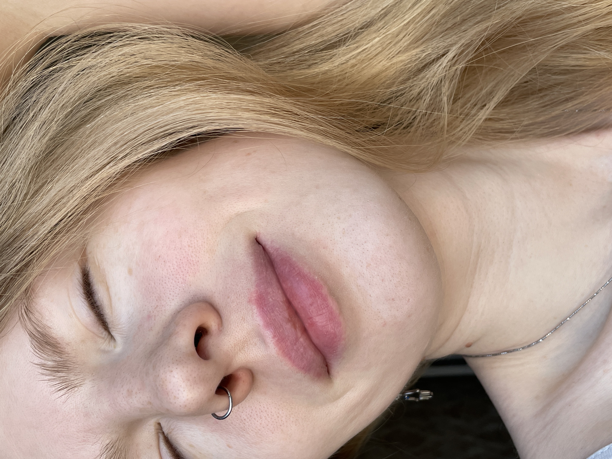 A close-up photograph depicts a fair-skinned individual, likely female, lying on their back. Her eyes are closed and her face is tilted to the right. She has light blonde hair, a nose ring in her left nostril, and her lips appear to be recently treated or swollen. A silver necklace is visible on her neck. The setting is non-descript and does not provide any indicators about being in Izhevsk, Russia.