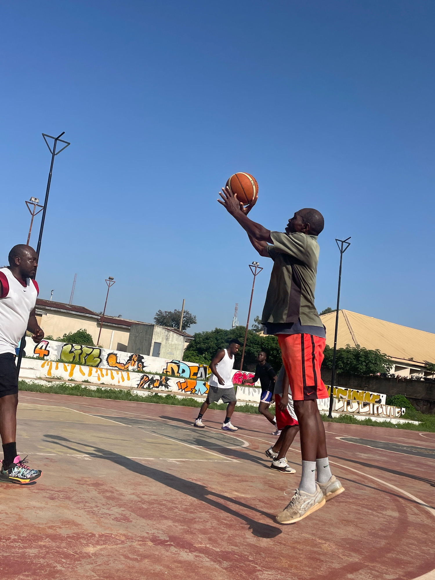 A group of men are playing basketball on an outdoor court. The court is made of concrete and has red and yellow markings. There are graffiti murals on a wall behind the court. The sky is clear and blue. The man in the foreground is wearing an olive green shirt and orange shorts, and he is jumping to shoot a basketball. He is bald and has dark skin. Another man in a white tank top and black shorts is on the left side of the court, watching. Two other men are in the background, one wearing a white tank top and gray shorts, and the other wearing a black t-shirt and blue shorts. The scene appears to be in Jos, Nigeria, given the presence of graffiti art common in the city and the overall environment.