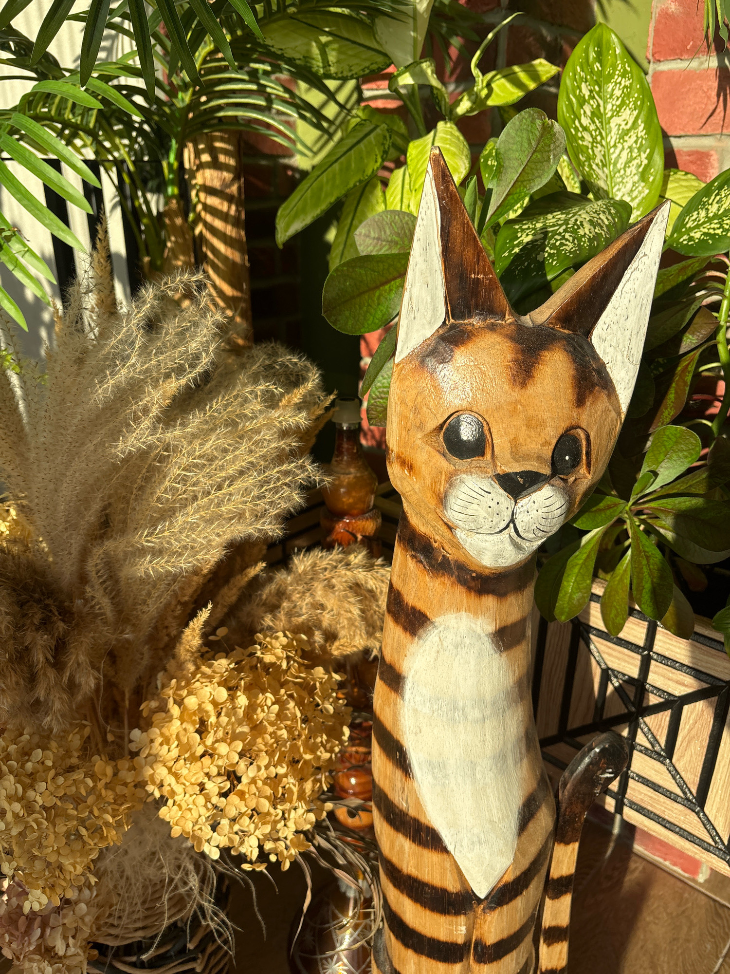 The image is a close-up, eye-level shot of a carved wooden cat figurine with orange and black stripes, white accents on its ears, chest, and face, and black painted eyes and whiskers. The cat is positioned in front of lush green foliage, a brick wall, and dried floral arrangements including pampas grass and hydrangeas. The lighting suggests an outdoor setting with strong sunlight casting shadows. The image does not provide any information about the city of Sapronovo, Russia.