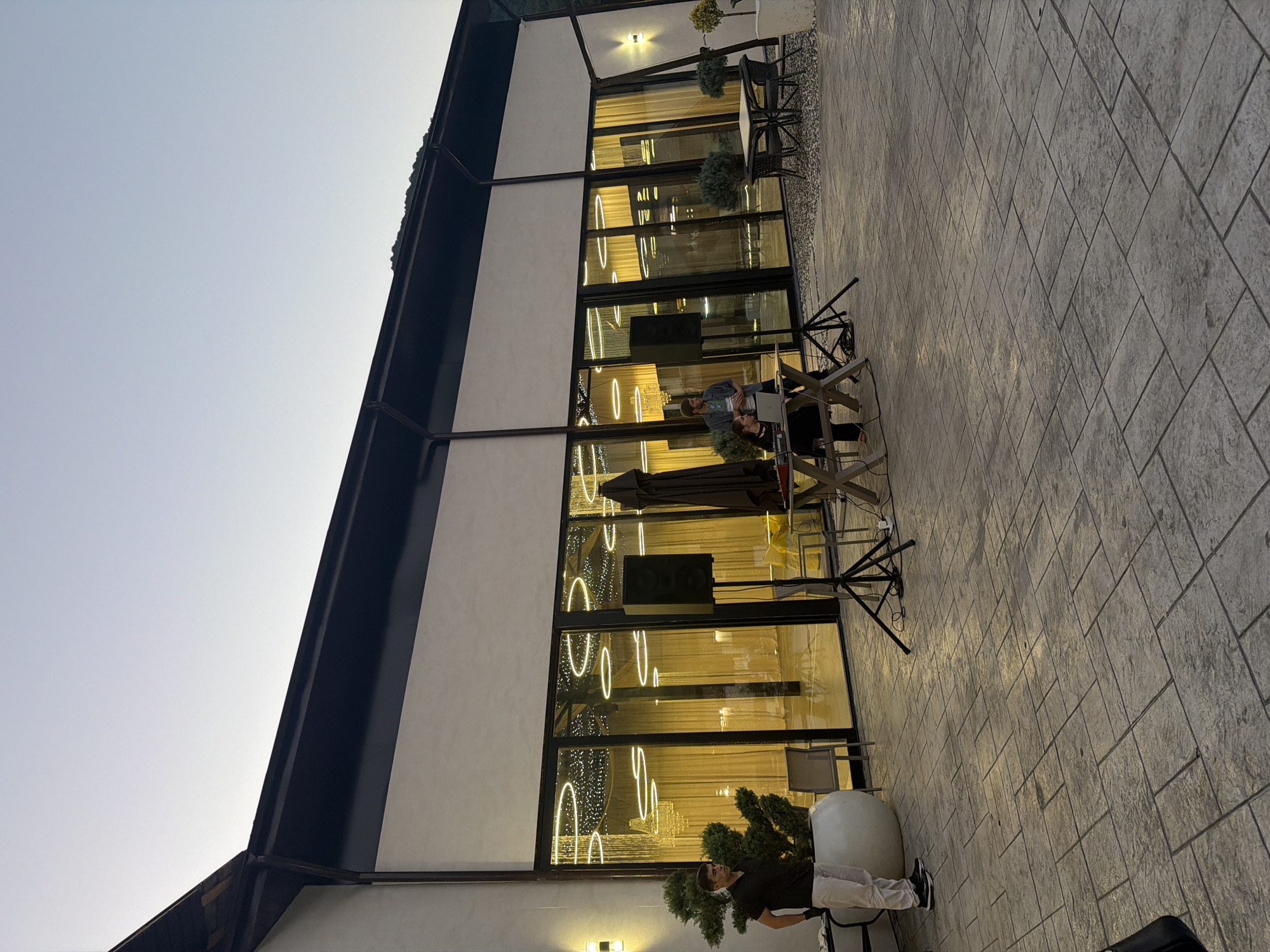 The image shows an outdoor patio area in front of a modern building with large glass windows. The building appears to be a venue, possibly for events, as indicated by the lighting and decor visible through the windows. The sky is a light, clear blue, suggesting it is either early morning or late evening.
In the foreground, a man in a black shirt and white pants stands near a large white planter containing a small evergreen tree. To his right, there are two large speakers on stands, positioned to face the building. Further to the right, there is a table with a laptop and some equipment, with two people standing behind it. One person appears to be operating the equipment, while the other is looking on. There are also several outdoor tables and chairs set up in the background, as well as some umbrellas. The patio is paved with large, rectangular stones.
The building's facade is white, with a dark-colored roofline. The large glass windows offer a view into the interior, which is decorated with circular light fixtures and what appear to be draped curtains or fabric. The overall impression is one of a contemporary and somewhat upscale event space. The location is identified as Achadara, Abkhazia, which places this scene in a specific geographical context.