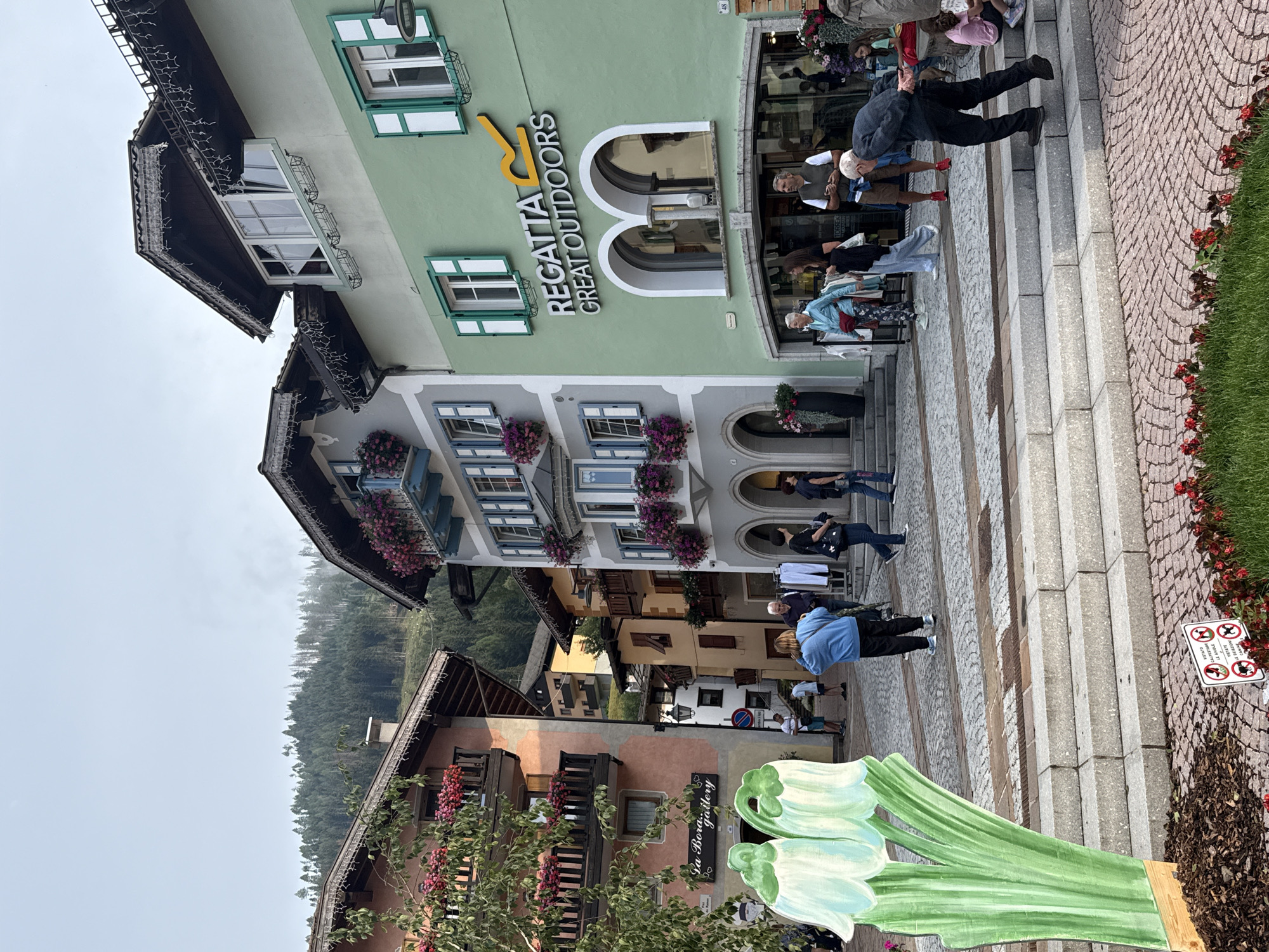The image depicts a street scene in Moena, Italy, a town nestled in the Dolomites. The architecture is characteristic of the region, with multi-story buildings featuring pitched roofs, balconies adorned with flowers, and shutters. One prominent building on the right is painted in a light green hue and displays the "REGATTA GREAT OUTDOORS" signage. In front of this store, several people are visible, some browsing merchandise displayed on racks outside. A man in a dark sweater and dark pants is walking down a set of stone steps, looking downwards. To the left, a building with a peach-colored facade features balconies with railings and more floral decorations. In the foreground, a large, colorful cutout resembling two green bell-shaped flowers stands on a wooden base. The street itself is paved with cobblestones and bordered by planters containing red flowers and greenery. The background shows a steep, tree-covered mountain slope under a hazy, overcast sky. The overall atmosphere suggests a tranquil day in a picturesque alpine town.