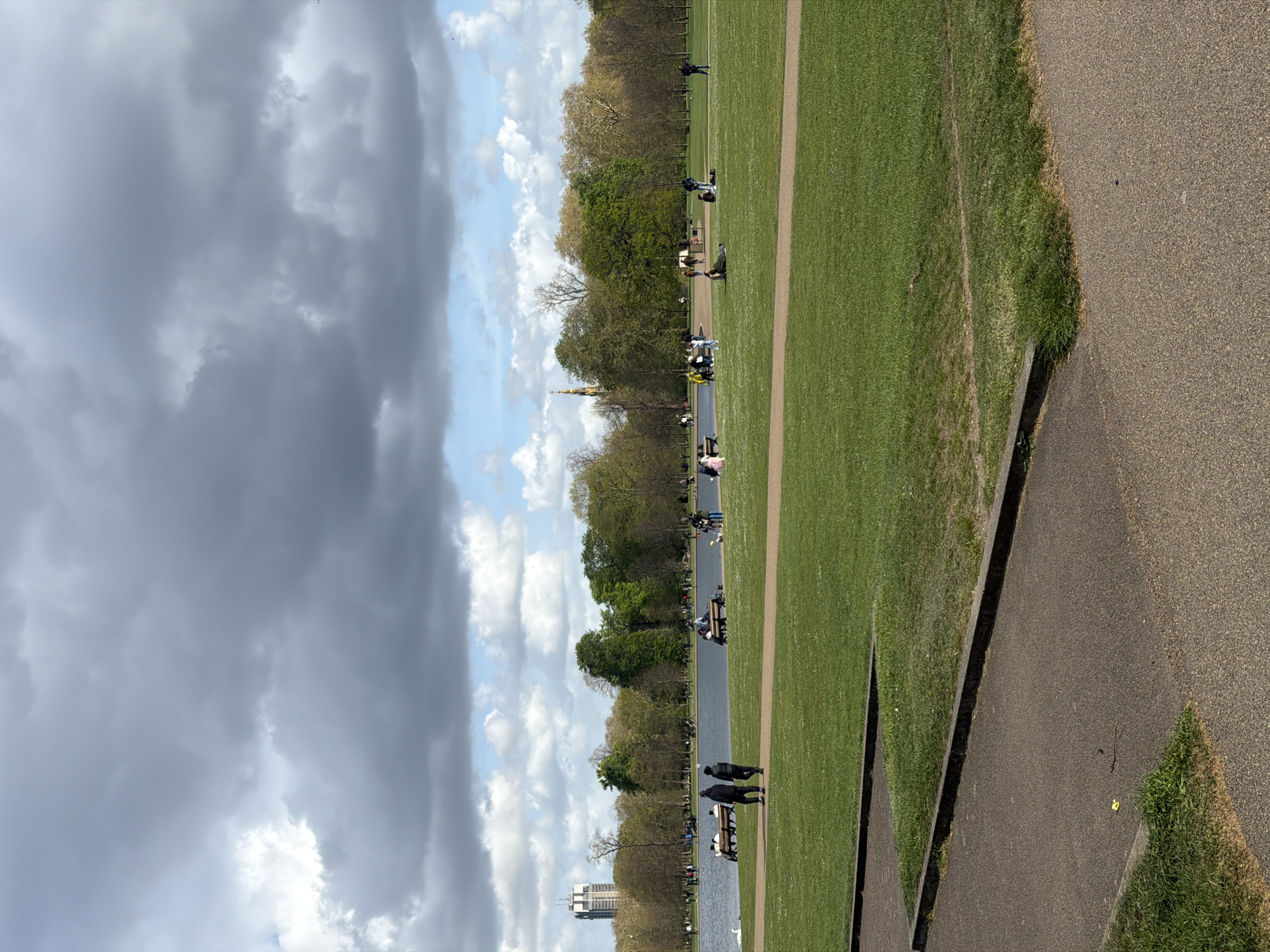 The image depicts a park scene in London, United Kingdom. A large expanse of green grass stretches across the middle of the frame, bordered by a paved pathway on the right and a line of trees and a body of water on the left. People are seen walking along the pathway and sitting on benches near the water. The sky is filled with a mix of dark, ominous clouds and patches of blue sky. In the distance, a tall building and the top of a monument are visible through the trees.