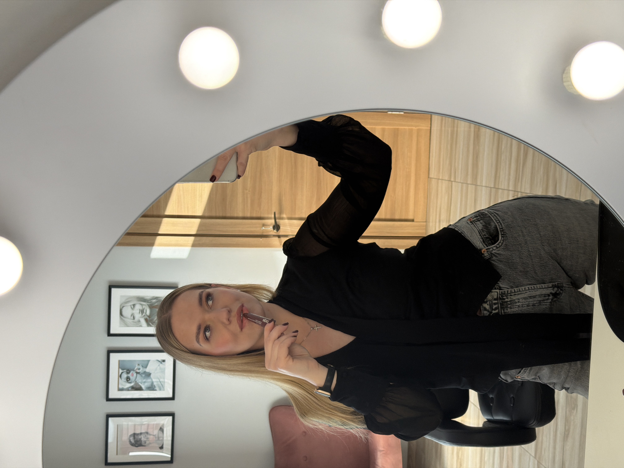A woman is reflected in a round, illuminated makeup mirror. She is holding a mobile phone in her right hand and applying lipstick with her left. She has long blonde hair, is wearing a black blouse, jeans, and a necklace. The reflection reveals a room with framed pictures on the wall, a pink chair, and a wooden door. The location is suggested to be Riga, Latvia, but is not visually verifiable.