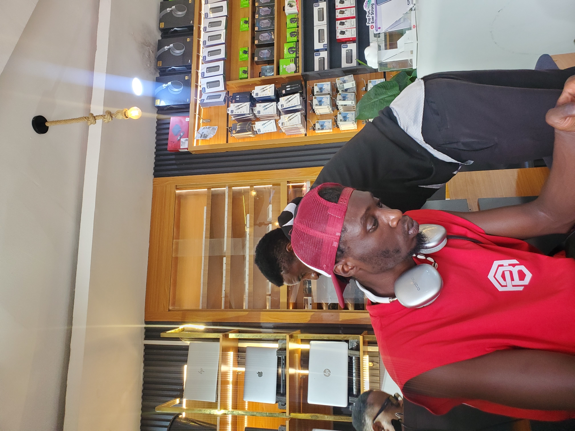 In a retail electronics store located in Jos, Nigeria, a young Black man wearing a red sleeveless t-shirt and a red cap is seated and looking down. He has large, silver headphones around his neck. To his right and behind him, another Black man in a black hoodie is leaning forward, seemingly interacting with something on a shelf. The store displays numerous electronic devices, including laptops on shelves to the left and rows of boxed accessories such as chargers, earphones, and power banks on shelves to the right. A rustic pendant light with a bare bulb hangs from the ceiling above. The environment appears to be well-lit, with artificial lighting from the ceiling and possibly from within the display shelves.