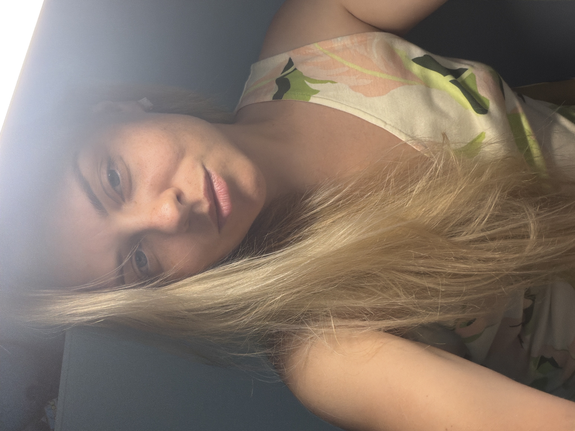 A medium close-up selfie shows a woman with blonde hair and fair skin. She is looking slightly off-camera, with her mouth slightly open. She is wearing a white tank top with a floral pattern in pink, green, and black. Her hair is long and wavy, falling over her left shoulder and obscuring part of her face. The background is dark and out of focus, with a bright light source above and to the right of her head, casting a strong glare on the upper part of the image. The woman appears to be indoors, possibly in a room with a dark wall. There is no identifiable information in the image that would indicate a specific location in Votkinsk, Russia.