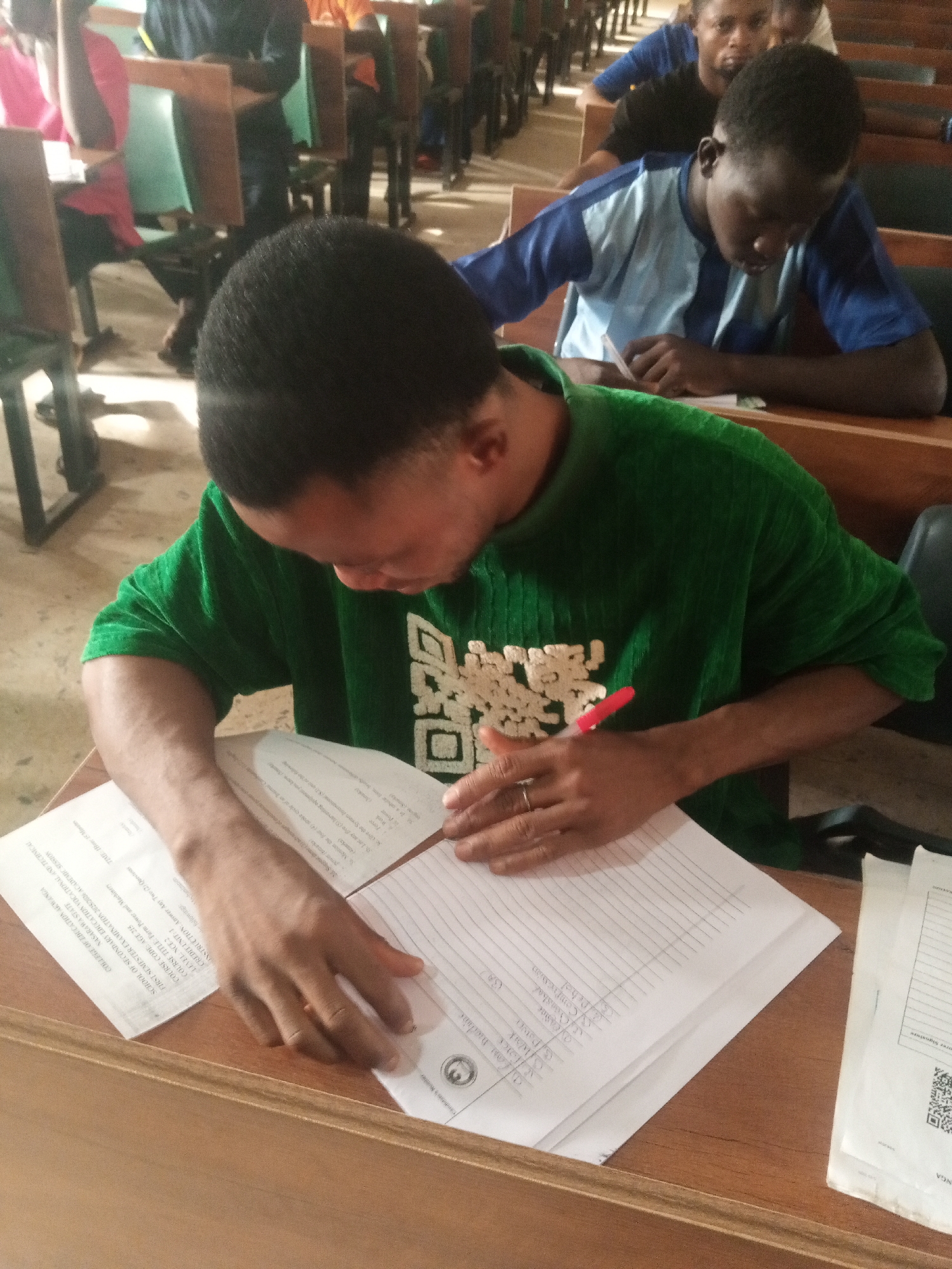 The image depicts an indoor scene within what appears to be a classroom or examination hall, bustling with students. In the foreground, a person wearing a green long-sleeved shirt with a light-colored patterned design is seated at a wooden desk, deeply engrossed in writing on paper with a red pen. A ring is visible on their left hand as they work.
Behind them, several other students are seated in rows of wooden desks. One student in the mid-ground, wearing a blue and light blue striped shirt, is clearly visible, also focused downwards, presumably on their own papers. The overall activity suggests an examination or a significant writing task.
Notable details include the various papers on the foreground desk, containing specific academic information. Visible text from these documents includes: "COLLEGE OF EDUCATION AKWANGA, NASARAWA STATE," "SCHOOL OF SECONDARY EDUCATION VOCATIONAL AND TECHNICAL," "FIRST SEMESTER EXAMINATION," "COURSE TITLE: Farm Power and Machinery," "COURSE CODE: AGE 213," "LEVEL: NCE2," and "CREDIT UNIT: 1." The instruction "Answer Any Two (2)" is also present, along with partial questions. Other discernible text snippets include "Patver," "Coashal," "Comprenin," "Pehol," "TIME: 1 Hour 40 Minutes," "Candidate's Number," and "Examiner's Signature," as well as a QR code on one of the sheets. The subjects appear focused and diligent. The lighting suggests it is daytime.