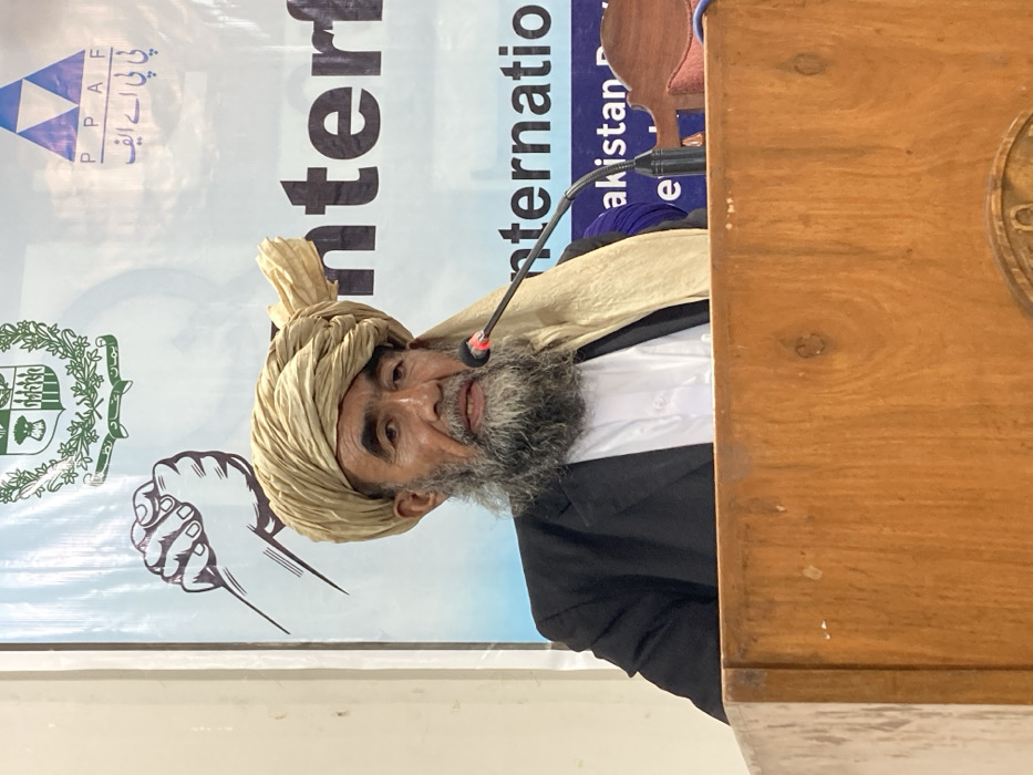 The image shows an elderly male subject speaking at a wooden podium in an indoor setting within Quetta City Tehsil, Pakistan. The man has a long, dark grey beard and mustache, and is wearing a light-colored (beige/khaki) turban, a dark suit jacket, and a white collared shirt. He is positioned centrally, with his mouth slightly open as if mid-speech, addressing an unseen audience. A black gooseneck microphone is extended from the podium towards his mouth.
Behind the speaker is a large banner, predominantly white and light blue. The top left of the banner displays a green emblem featuring trees, a crescent moon, and a star, with Urdu text below it identifying "حکومت بلوچستان" (Government of Balochistan) and "محکمہ سماجی بہبود" (Department of Social Welfare). On the top right, a blue triangular logo for "PPAF" (Pakistan Poverty Alleviation Fund) is visible, accompanied by its Urdu translation. Below the Balochistan emblem, an illustration of two clasped hands in a handshake is present. Large, black English text partially reads "Interf" and "Internatio," suggesting an event on interfaith or international dialogue, with additional smaller English text "akistan D" visible below. The wooden podium in front of the speaker has visible wood grain and a partially obscured carved emblem at its lower front. The immediate background wall to the left of the banner is a light, neutral color.