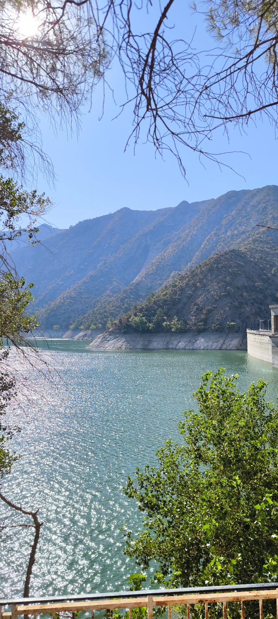 The image shows a reservoir in the municipality of Peramola, Spain, under a clear blue sky. The reservoir's surface is a vibrant turquoise, reflecting the sunlight with a dazzling shimmer. The water is calm, with gentle ripples breaking the surface. Along the shoreline, a rugged, rocky embankment slopes down to the water's edge. Further in the background, a series of majestic, tree-covered mountains rise, their slopes a mix of green vegetation and rocky outcrops. The mountains appear layered, fading into a hazy blue as they recede into the distance. On the right side of the frame, a portion of a concrete dam structure is visible, its imposing presence indicating the reservoir's function as a water storage facility. In the foreground, branches of trees frame the scene, their leaves a lush green, adding a natural border to the expansive view. A metal railing runs along the bottom of the frame, suggesting a viewing platform or walkway overlooking the reservoir. The overall scene evokes a sense of natural beauty and impressive engineering, highlighting the picturesque landscape of Peramola.