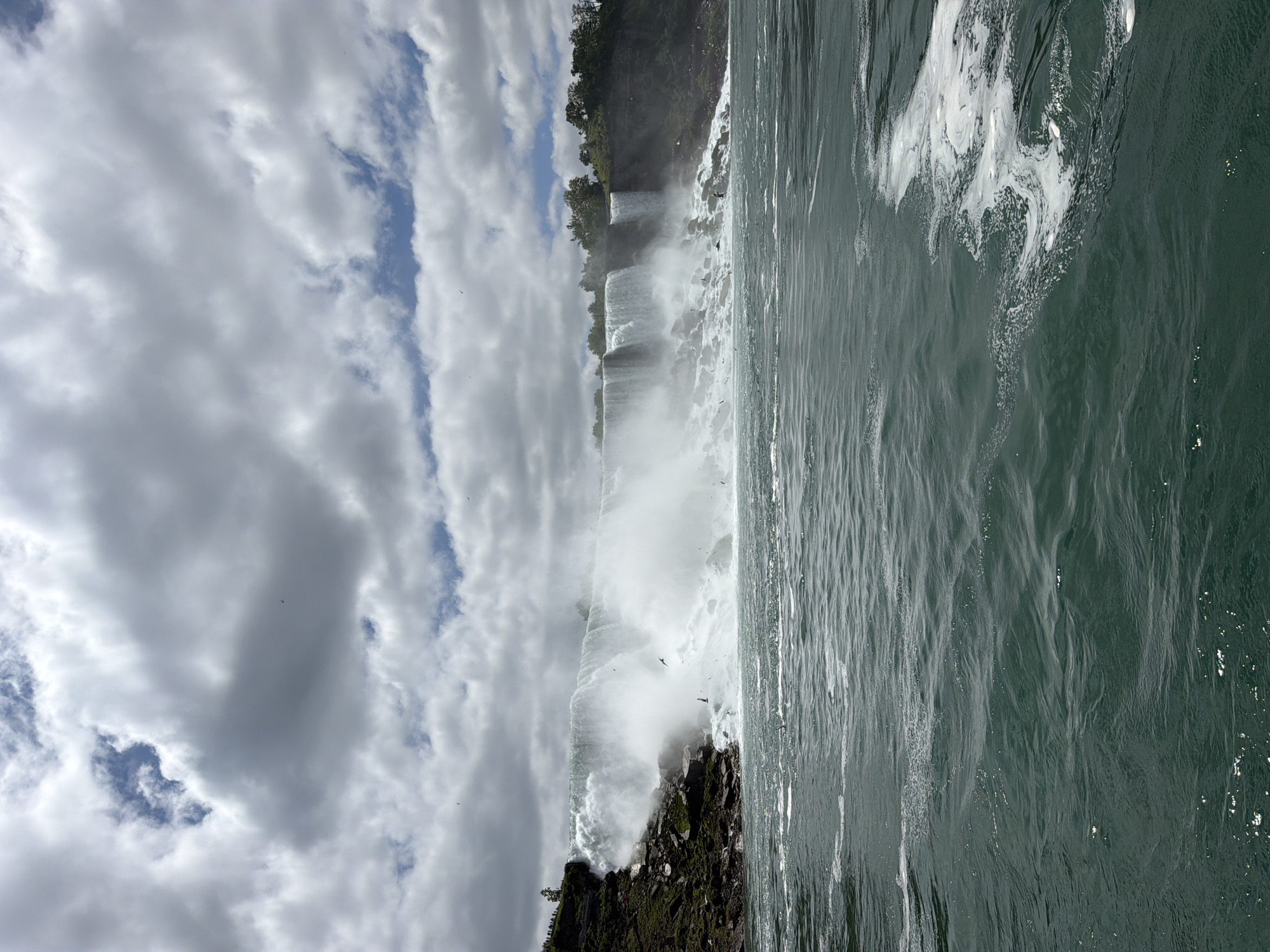 This image captures a portion of Niagara Falls, specifically the American Falls and Bridal Veil Falls, viewed from the Niagara River in the City of Niagara Falls, United States of America. The perspective is low, suggesting observation from the water level.
The upper third of the image shows a cloudy sky, dominated by large, uneven gray and white clouds. Patches of blue sky are visible through breaks in the cloud cover, particularly in the upper left and right quadrants. Several small, dark avian figures are scattered across the sky and near the falls.
The mid-ground is dominated by the cascading water of Niagara Falls. To the left, a large volume of white, turbulent water descends over a dark, rocky cliff face. A dense mist rises vigorously from the base of the falls, partially obscuring the lower portions of the falling water and the rock formations. On the upper left bank, above the falls, a small cluster of indistinct human figures is visible. The cliff face itself is rugged, composed of dark rock and sparse green vegetation. Further to the right, additional segments of the waterfall are visible, flowing over a less steep, vegetated embankment. Green trees line the upper edge of the cliff across the visible expanse of the falls. Numerous small, dark birds are actively flying in the mist and turbulent air directly in front of the waterfall.
The lower half of the image consists of the dark, deep green water of the Niagara River. The water surface is rippled and shows considerable movement, with areas of white foam and agitation, particularly in the mid-right and closer to the base of the falls. Scattered glints of light are reflected on the water's surface in the foreground.