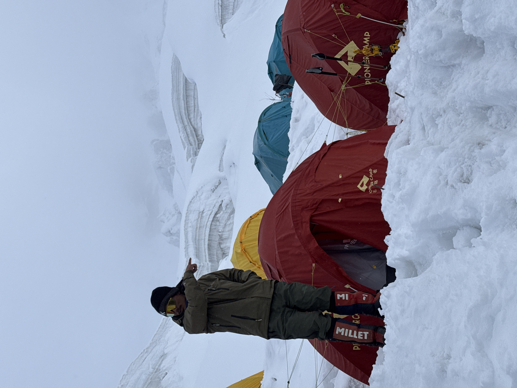 A person wearing a green jacket, green pants, a black beanie, and sunglasses stands in the snow, pointing to the right. They are wearing red boots with white lettering that says "MILLET" and "MI." In the background, there are several tents, including red, yellow, and blue ones, set up on a snowy mountainside. The environment appears to be a high-altitude glacial area, possibly part of a base camp. The sky is overcast and hazy, with snow-covered peaks visible in the distance. The overall scene suggests an expedition or climbing activity in a remote, cold environment, potentially near Samagaun, Nepal, given the context of Himalayan mountaineering.