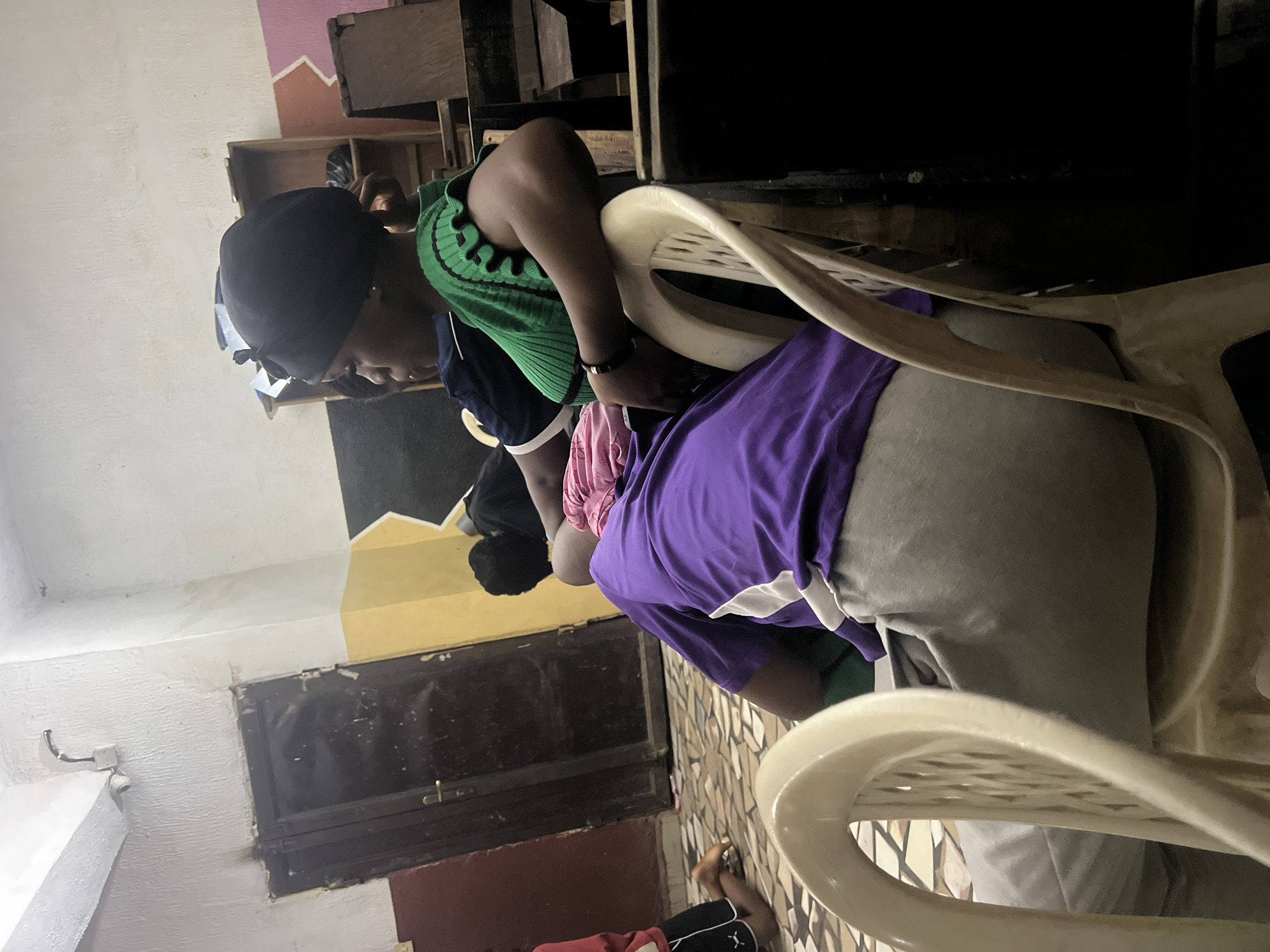 In Nkpor, Nigeria, an indoor scene captures several individuals in what appears to be a communal space. In the foreground, a person sits in a plastic chair, draped in a purple garment and gray pants. To their left, another individual wears a green ribbed top and a black head covering, leaning back and looking to the side. Behind them, another person is visible, their head bowed. The room features a closed, dark double door on the left, with a security camera mounted above it on a textured white wall. Yellow and dark gray paint adorns the wall further back. Wooden shelving units and dark furniture are present in the background, with a tiled floor completing the environment.