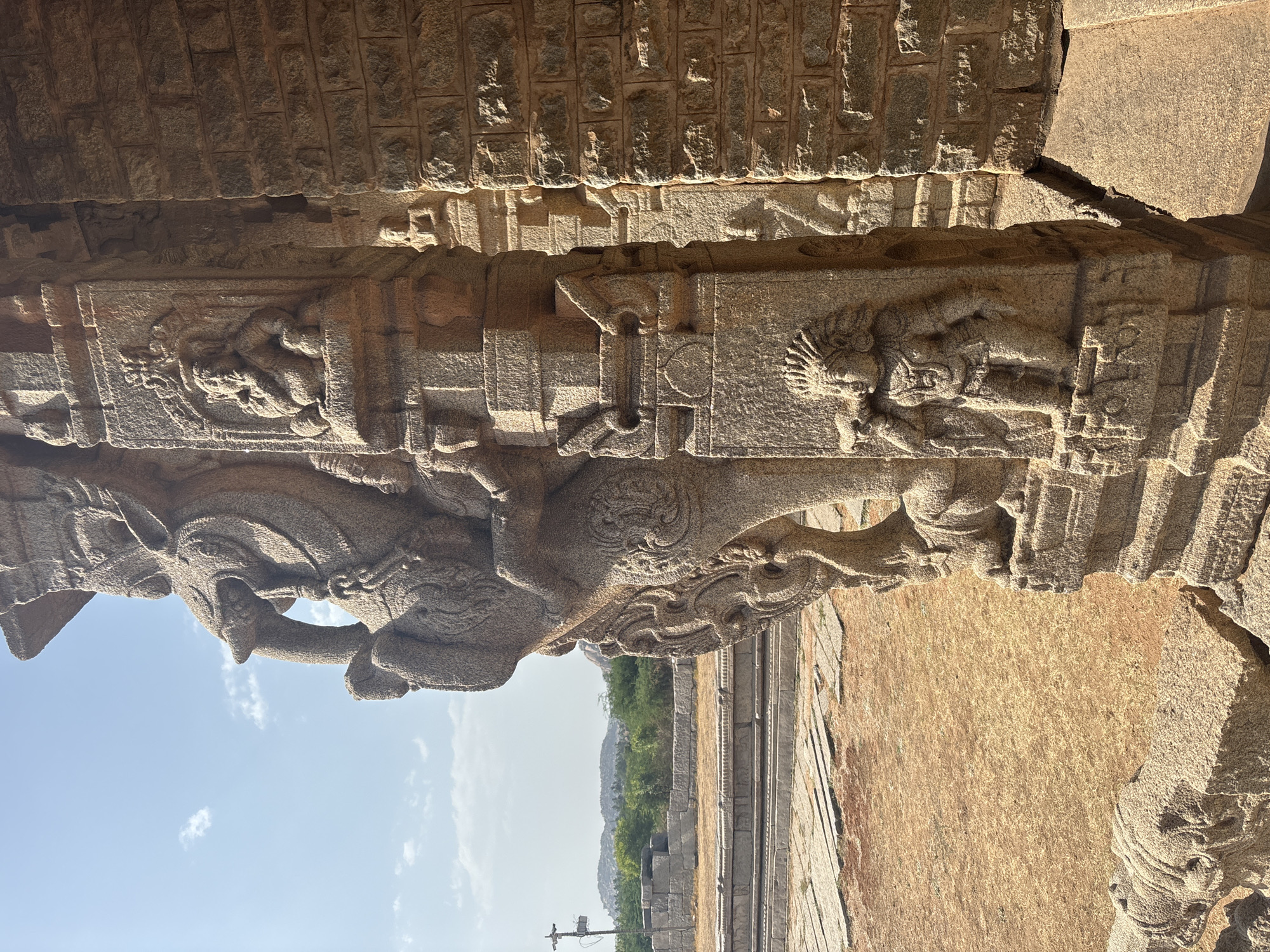 The image showcases intricate stone carvings on a structure in Hampi, India. The carvings feature human and animal figures, possibly depicting mythological scenes. The stone architecture is weathered, indicating age and exposure to the elements. In the background, a landscape with vegetation is visible under a clear sky. The structure's design and material are indicative of the region's historical and architectural heritage.