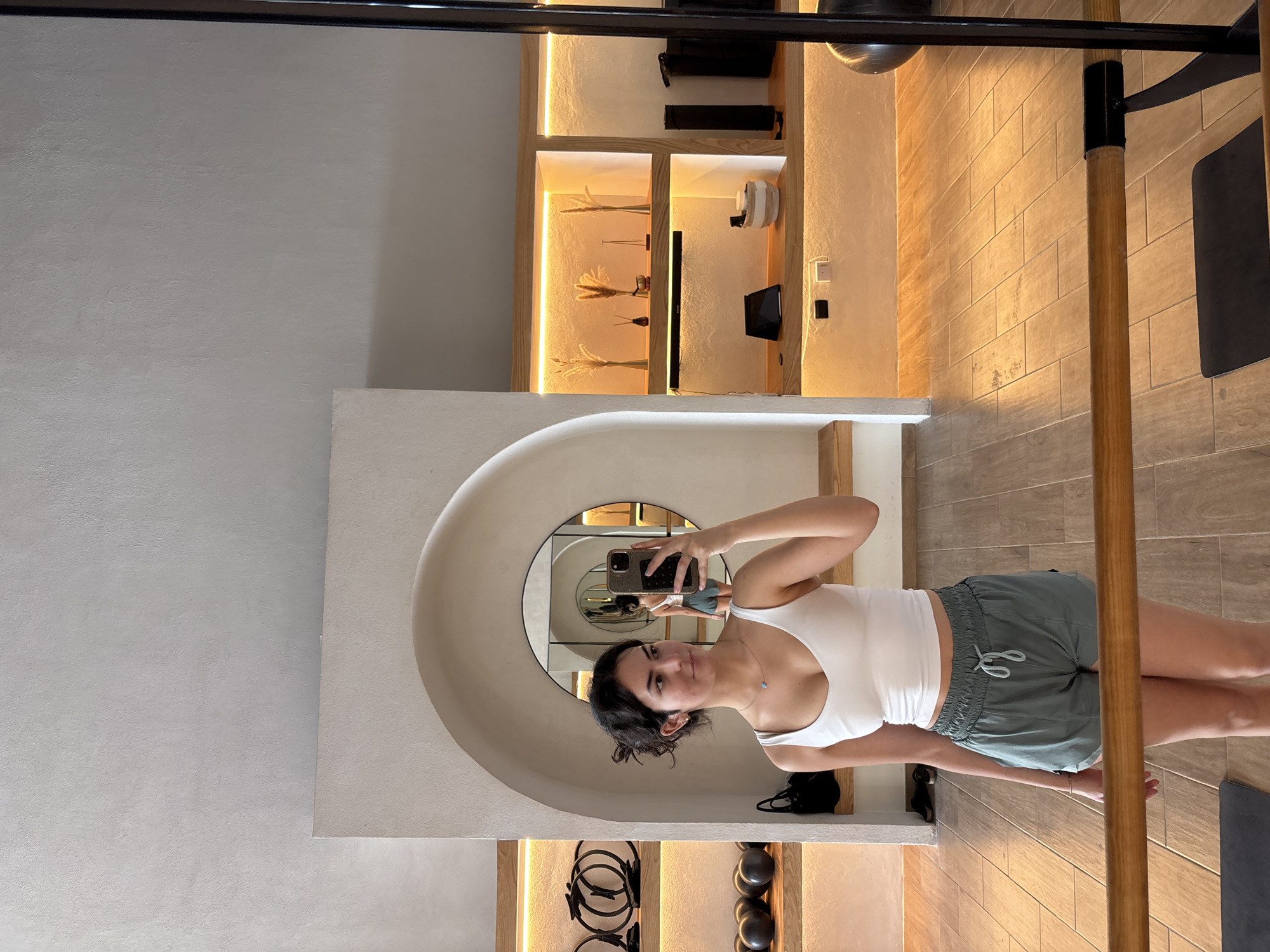 A woman is taking a selfie in a mirror at a fitness studio. The mirror is an oval shape and the woman is standing in front of it, holding her phone up to take the picture. She is wearing a white tank top and green shorts. The fitness studio has light wood floors and white walls. There are shelves with workout equipment on them in the background. The woman is in San Bernardino Tlaxcalancingo, Mexico.