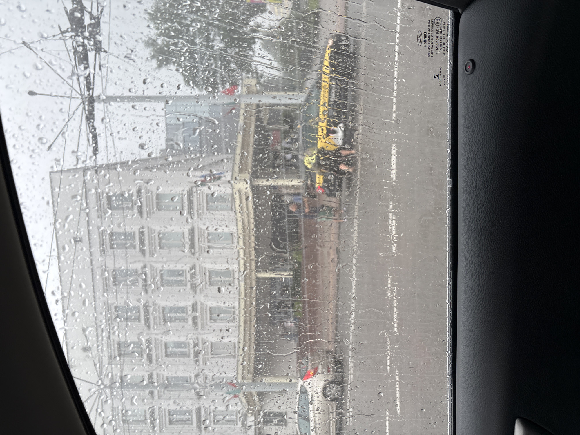 This image depicts a view from inside a vehicle, looking through a rain-streaked window at a street scene in Vitebsk, Belarus. The building in the background appears to be a large, white, multi-story structure with many windows, possibly a hotel or administrative building, with an awning or covered walkway in front. Traffic lights and overhead wires are visible, suggesting a busy urban environment. A white car is parked on the left side of the street, and a yellow taxi with a person leaning into its open door is on the right. Several other people are present, though their actions are indistinct due to the rain and distance. The road surface is wet and reflective, indicating recent or ongoing rainfall. The foreground of the image shows the dark interior of the vehicle, including part of the door panel. A small red indicator light is visible on the door panel. A sticker with text and logos is affixed to the lower right corner of the window. The overall impression is one of a gray, rainy day in a European city.