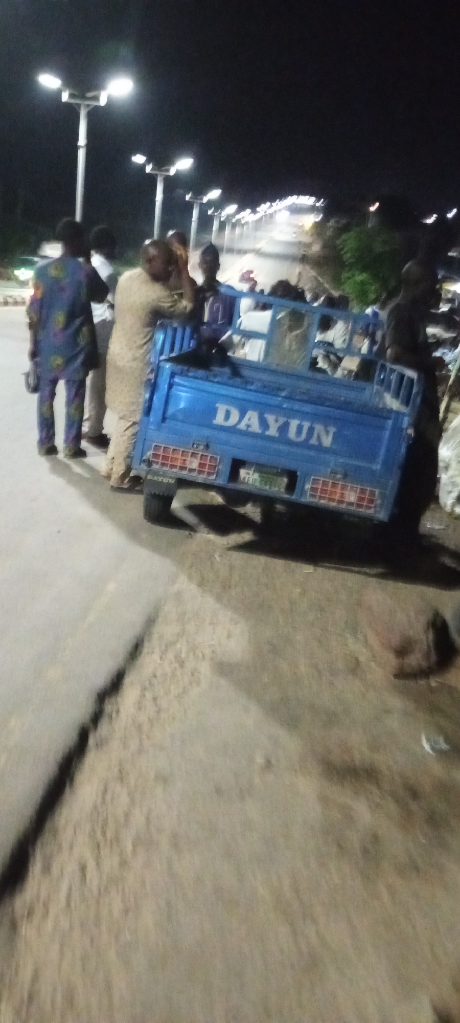 In Adankolo, Nigeria, a nighttime scene unfolds with several individuals gathered around a blue three-wheeled vehicle. The vehicle, a Dayun truck, has its name prominently displayed on the side. The surrounding environment appears to be a road or pathway, illuminated by streetlights that cast a glow on the scene. Some of the individuals are standing near the truck, while others are in the process of loading or unloading items. The overall atmosphere suggests a functional or logistical activity taking place after dark.
