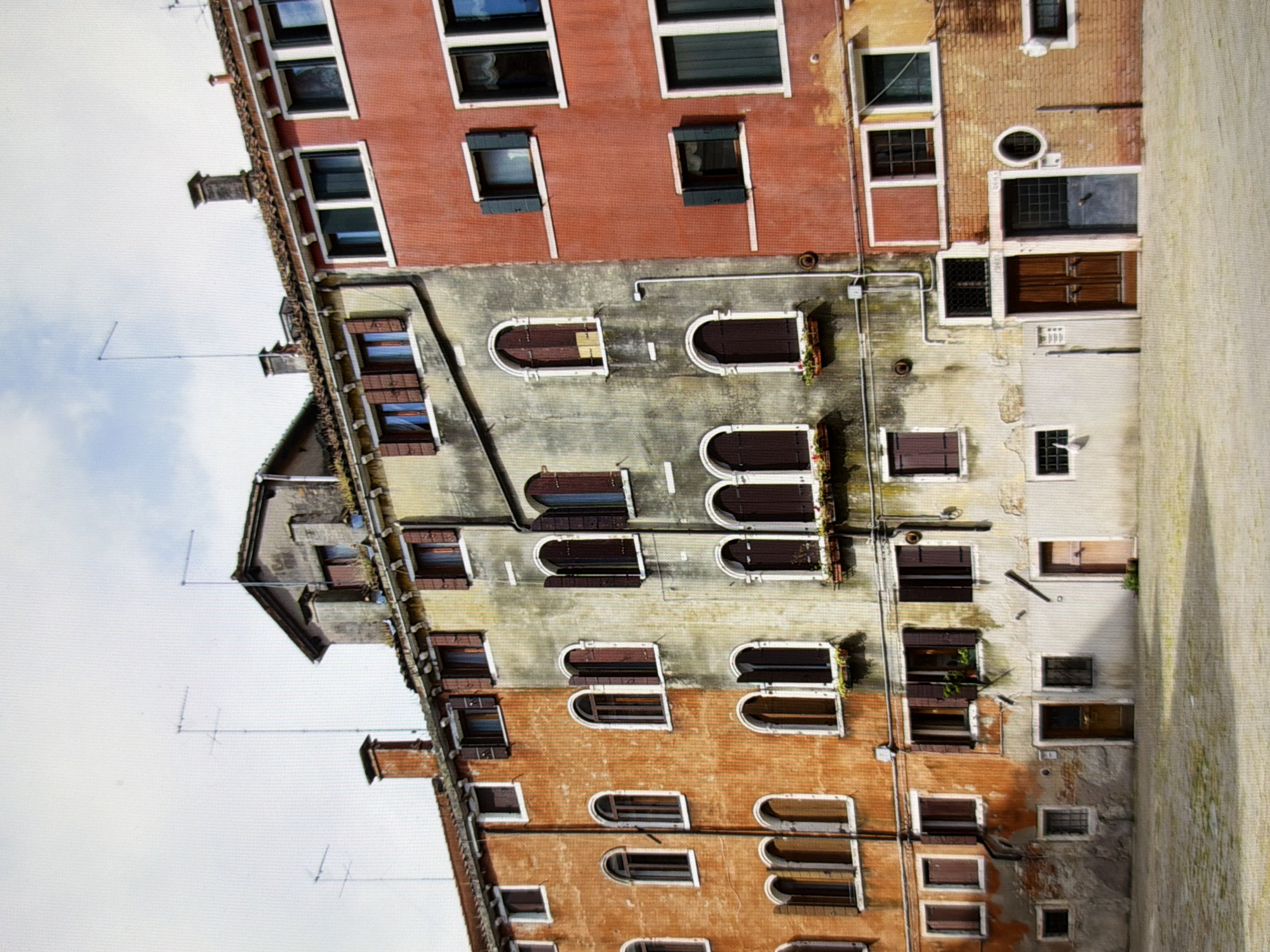 This is a medium-shot, eye-level, outdoor, wide-angle photograph of a row of old, multi-story buildings. The buildings are constructed of brick and stucco, with terracotta tiled roofs. The stucco is weathered and peeling in places, with a mix of orange, yellow, and grey tones. The buildings have numerous windows, many with dark brown wooden shutters, some of which are open. There are also arched windows on the lower floors, and some windows have flower boxes. Several chimneys are visible on the rooftops, along with modern television antennas. A paved courtyard with a textured surface is in the foreground, and the sky above is partly cloudy. The image is taken in Venice, Italy.