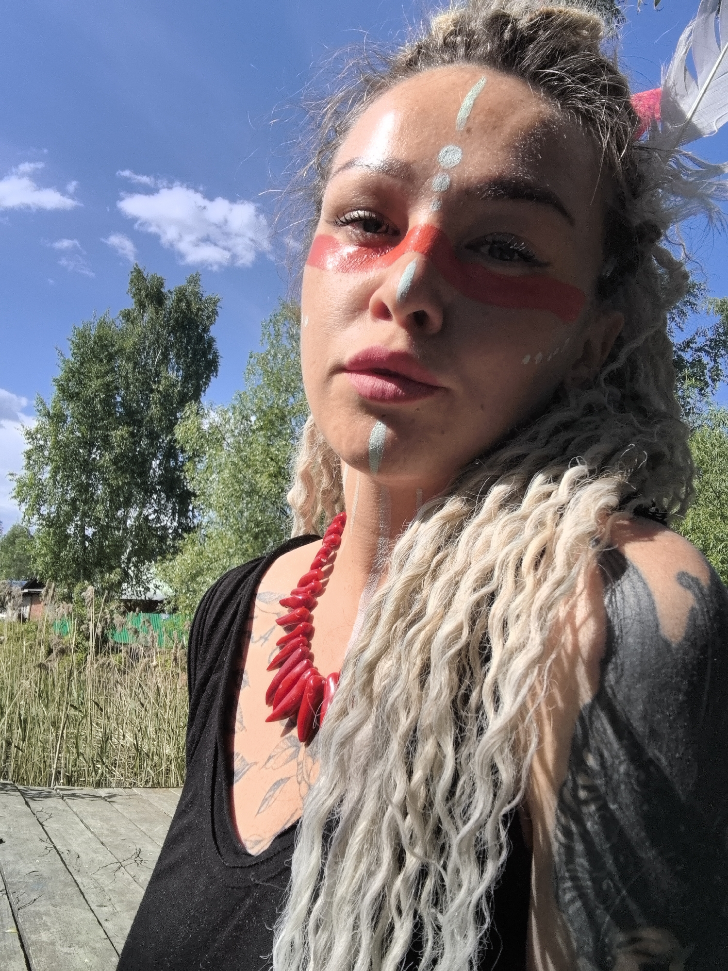 The image is a selfie of a woman with light complexion and blonde dreadlocks, wearing red and white face paint markings, a large red necklace, and a black shirt. A feather is visible in her hair. The background includes a blue sky with some clouds, trees, and a wooden platform. A building with a green corrugated metal roof is also visible amidst the trees. The location appears to be outdoors near a body of water. The city is not mentioned.