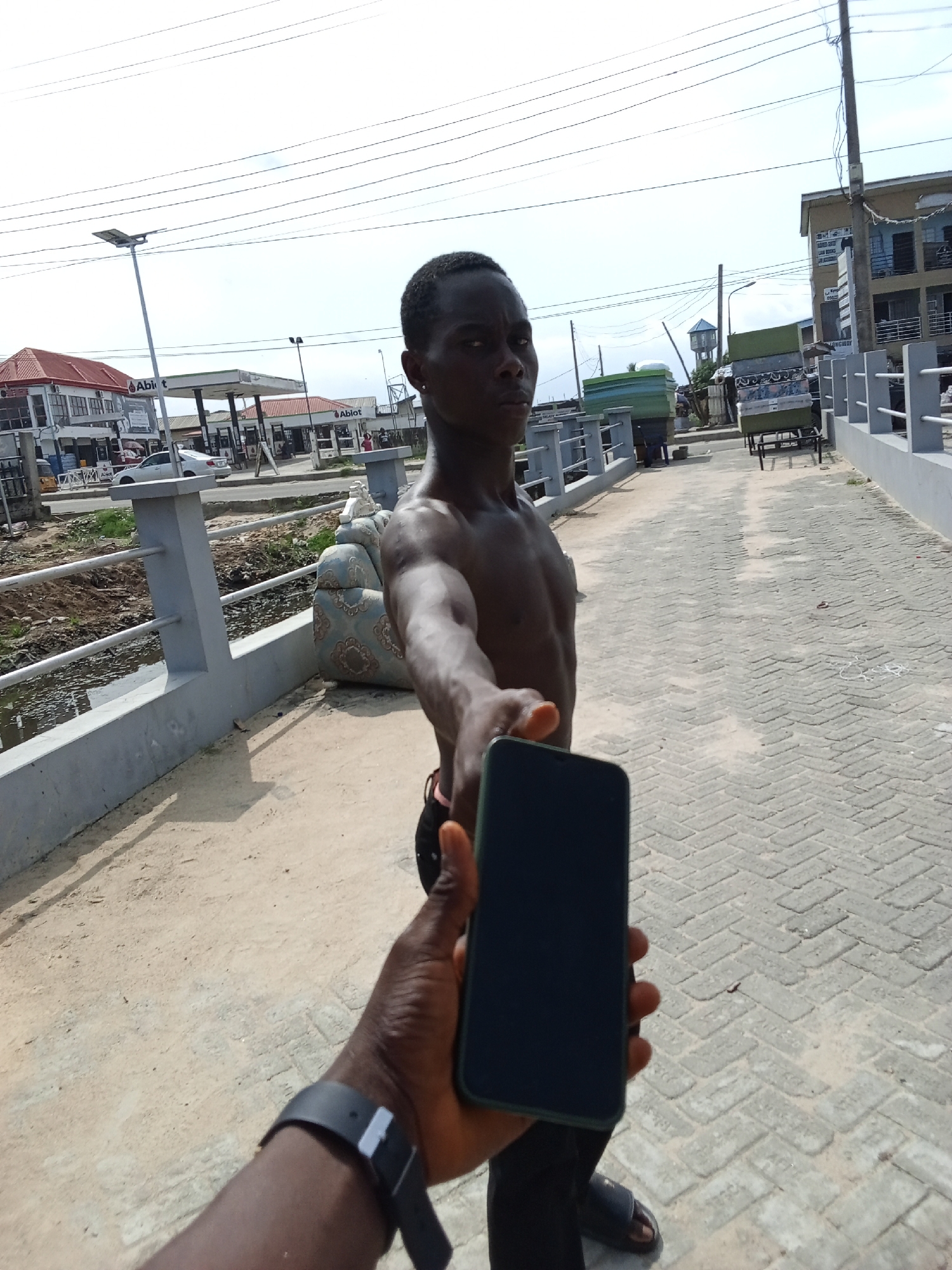 This medium-shot photograph, taken outdoors in bright daylight, captures a street scene likely in Ado, Nigeria. The perspective is from a person holding a dark-colored smartphone, whose arm, hand, and a dark smartwatch are visible in the immediate foreground.
In the mid-ground, a young adult male with dark skin is the primary subject. He is shirtless, wearing dark pants or shorts and dark sandals, and looks directly at the camera with a neutral to serious expression. He appears to be extending his right arm forward towards the camera.
The background features a paved pathway constructed from light-colored interlocking bricks, with concrete railings on both sides. To the left of the path, beyond the railing, is a ditch or canal with dirty water and some debris. A discarded blue and gold patterned armchair is leaning against the left railing. Further left, a petrol station with red roofs and signs partially visible as "Ablot" or "Abit" stands, with a white car parked nearby.
Overhead, multiple power lines stretch across a bright, clear sky. A tall street light with a solar panel is visible on the left, and a water tower is in the distant background. On the right side of the path, past the railing, a multi-story building can be seen. Further down the path, two pushcarts, loaded with what appear to be mattresses or large wrapped items, are visible. The bright sunlight and minimal shadows suggest the photo was taken during midday or early afternoon.
