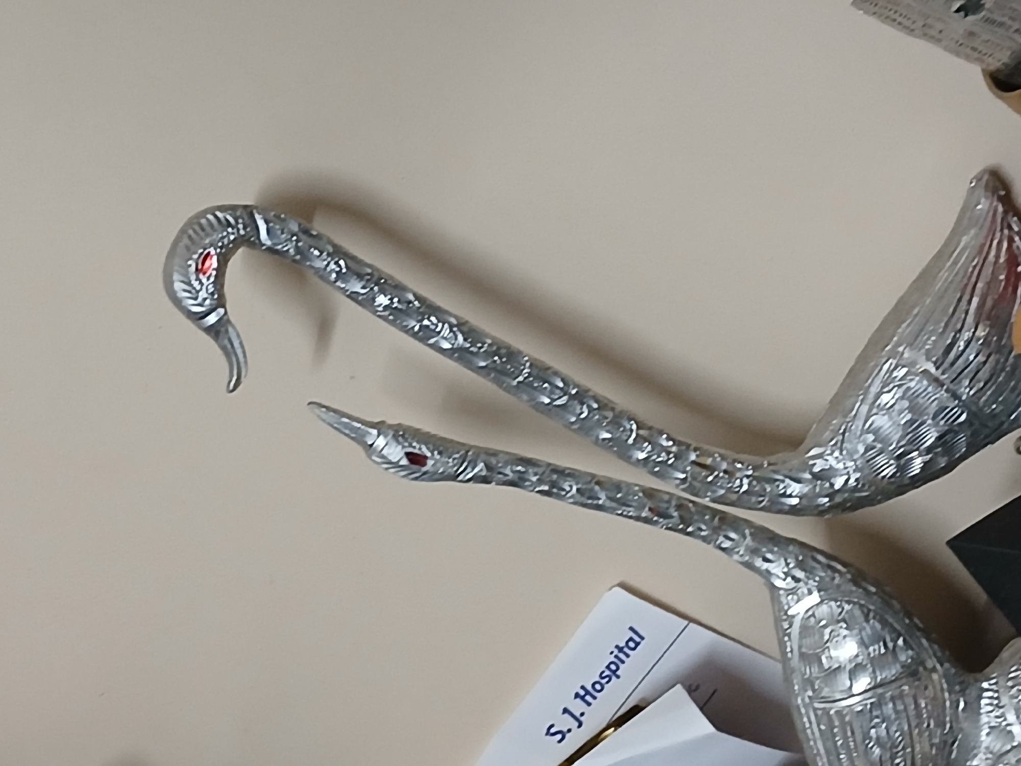 The image captures two ornate, metallic swan or flamingo-like decorative figures resting on a light-colored, plain surface, likely a tabletop. These figures are crafted from a shiny, silver-toned material, possibly chrome or silver plating, and exhibit intricate textures resembling scales or stylized feathers along their necks and partially visible bodies. Each figure features a slender, graceful neck that curves into a bird-like head with a pointed beak and a distinct red "eye."
The scene is an indoor setting, and no activity is taking place; the objects are static. In the lower part of the image, beneath one of the metallic figures, a white piece of paper is visible. Clear blue text on this paper reads "S.J. Hospital," providing a key location cue, suggesting the setting is associated with a hospital in Bengaluru, India. Indistinct crumpled paper or packaging material is partially visible in the upper right corner, and a dark, unidentified object can be seen in the bottom right. The lighting is even, illuminating the reflective surfaces of the figures and casting soft shadows, with no specific time of day or weather discernible.