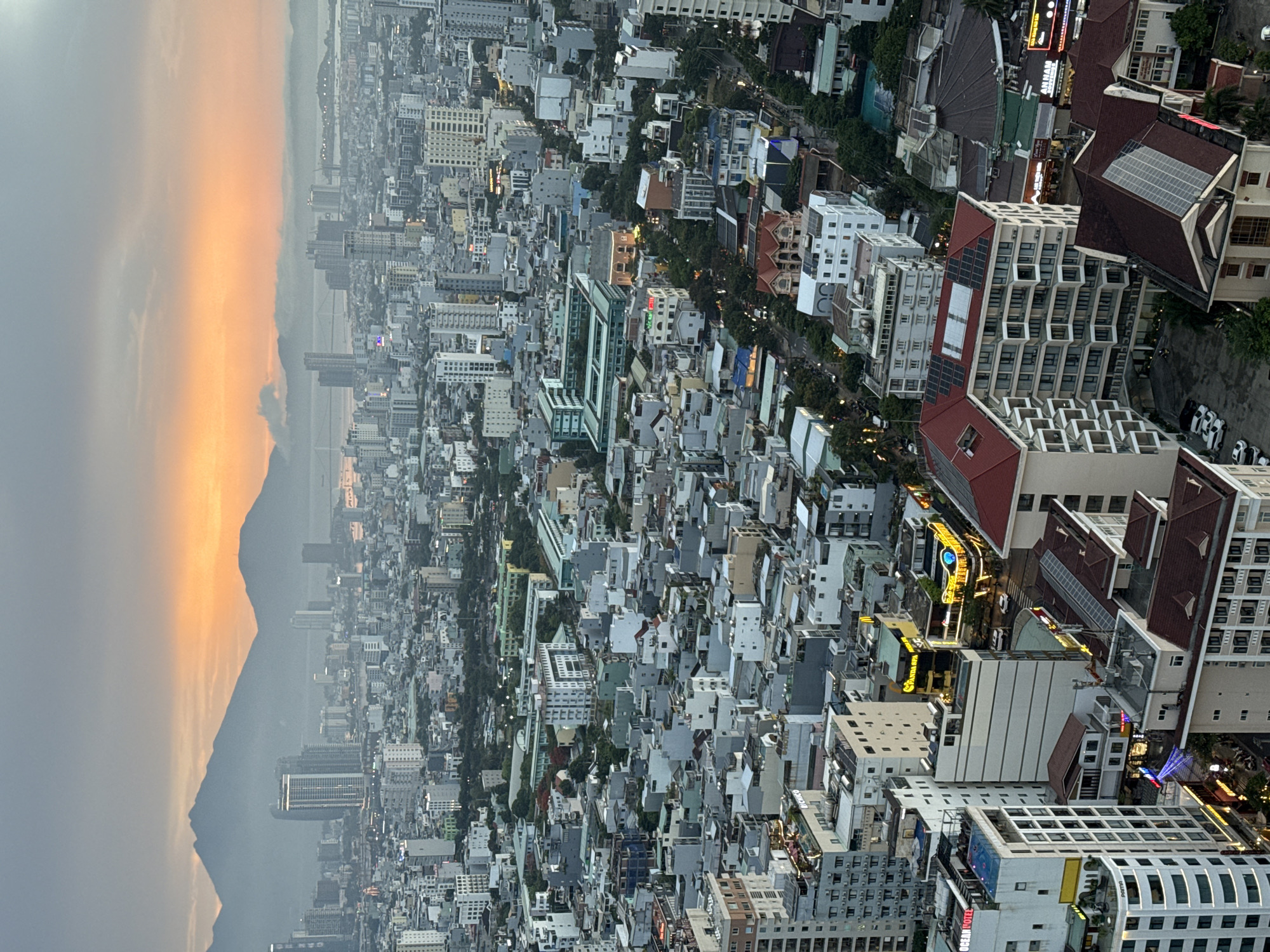 The image is a high-angle, full-frame shot of the city of Da Nang, Vietnam, during twilight. The sky is dominated by a gradient of grey clouds transitioning to a vibrant orange and yellow sunset glow towards the horizon, where mountains are silhouetted. The city below is a dense urban landscape filled with numerous buildings of varying heights and architectural styles, primarily constructed from concrete and glass. Many of these buildings are illuminated, suggesting it is evening.
In the foreground and midground, a variety of structures are visible, including multi-story apartment buildings, hotels, and commercial establishments. Some buildings have distinctive features like balconies, rooftop swimming pools, and solar panels. Several signs in Vietnamese are discernible on some of the buildings, indicating commercial activity. The streets below are visible in sections, with a few cars and motorbikes present. Trees and green spaces are interspersed throughout the urban sprawl.
In the background, the mountains form a dramatic backdrop against the colorful sky. A body of water, likely the East Sea, is visible in the distance, with a bridge crossing it. The overall impression is a bustling coastal city at dusk, with a blend of modern architecture and natural scenery. There are no discernible people or specific actions taking place that would suggest a narrative. The scene is purely an observational depiction of the urban environment.