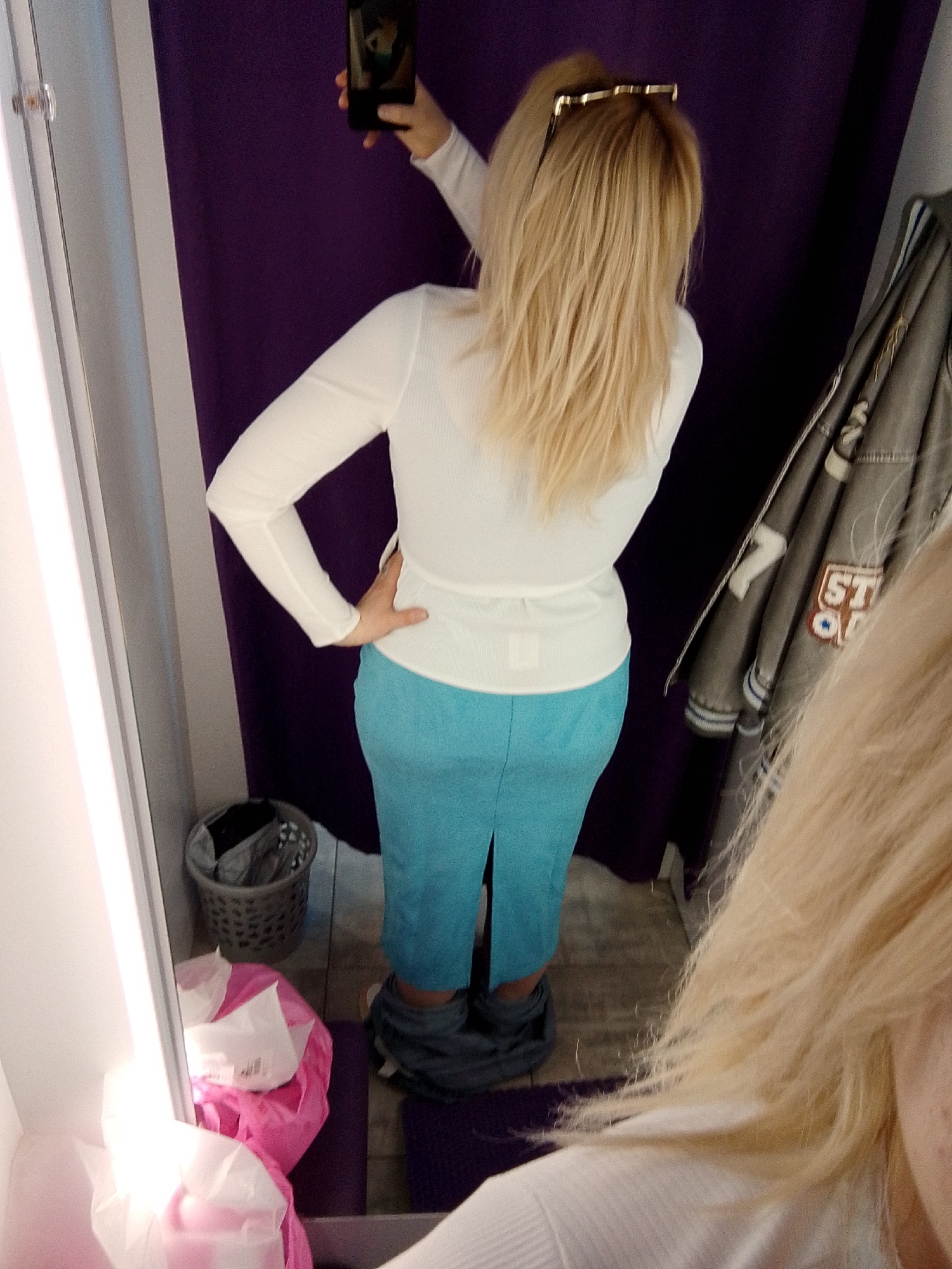 The image depicts a blonde woman taking a selfie in what appears to be a dressing room. She is wearing a white long-sleeved shirt and a blue skirt. Her pants are around her ankles. A jacket hangs on the wall next to her. The dressing room has a purple curtain. A trash can and shopping bags are on the floor. There is no definitive information in the image to confirm the city location in Izhevsk, Russia.