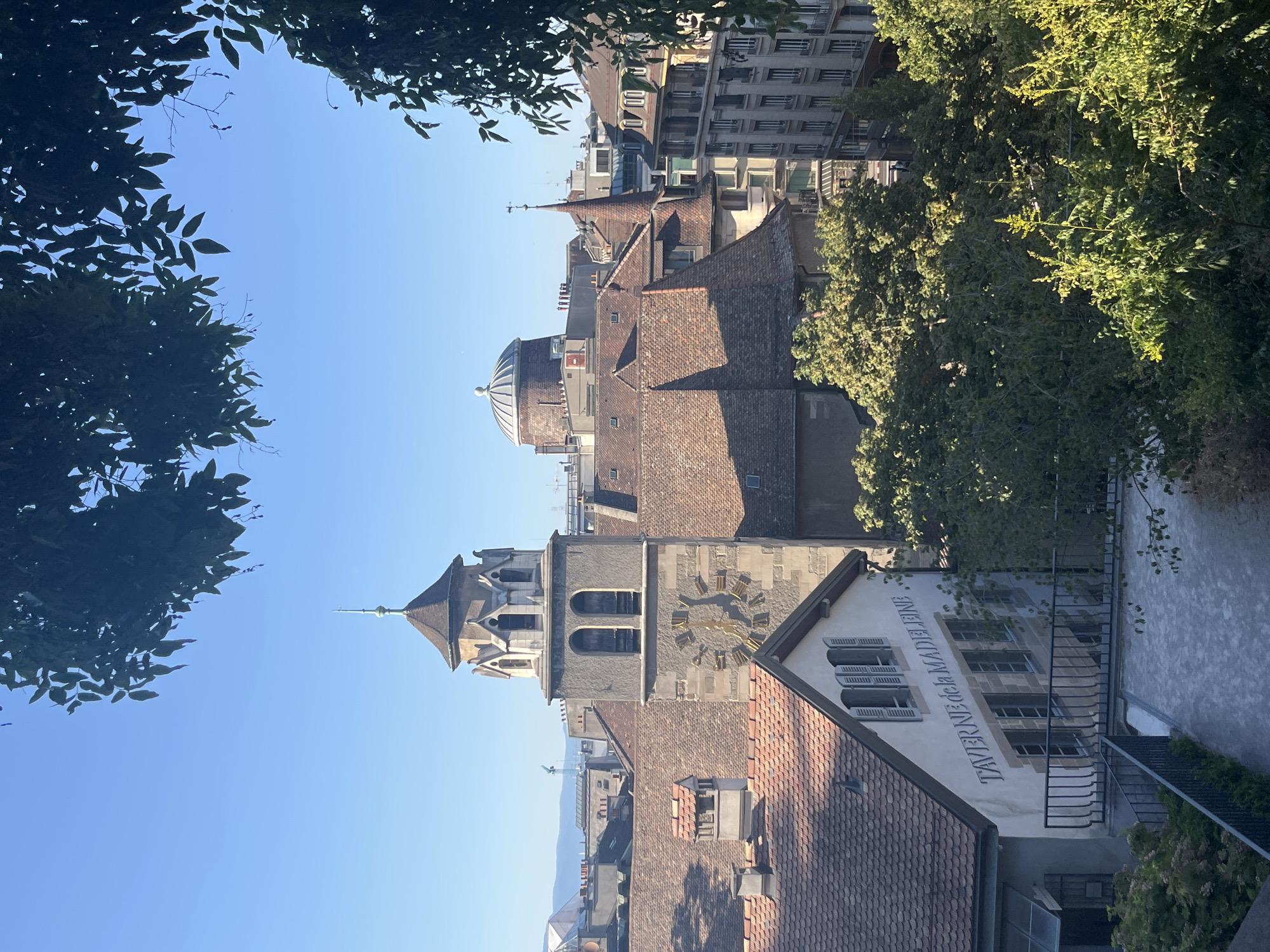 The image shows a cityscape in Geneva, Switzerland, with a clear blue sky and lush green trees in the foreground and mid-ground. In the center, a tall clock tower with a spire stands prominently, featuring a large clock face with Roman numerals. Adjacent to the tower, terracotta-tiled roofs of various buildings are visible, some with spires and dormer windows. To the left of the clock tower, a white building with the inscription "TAVERNE de la MADELEINE" is identifiable, featuring multiple windows and shutters. In the background, more buildings of varying architectural styles line the horizon, and distant hills or mountains are faintly visible. Sunlight casts shadows across the scene, indicating it is daytime. No people are visible in the image.
