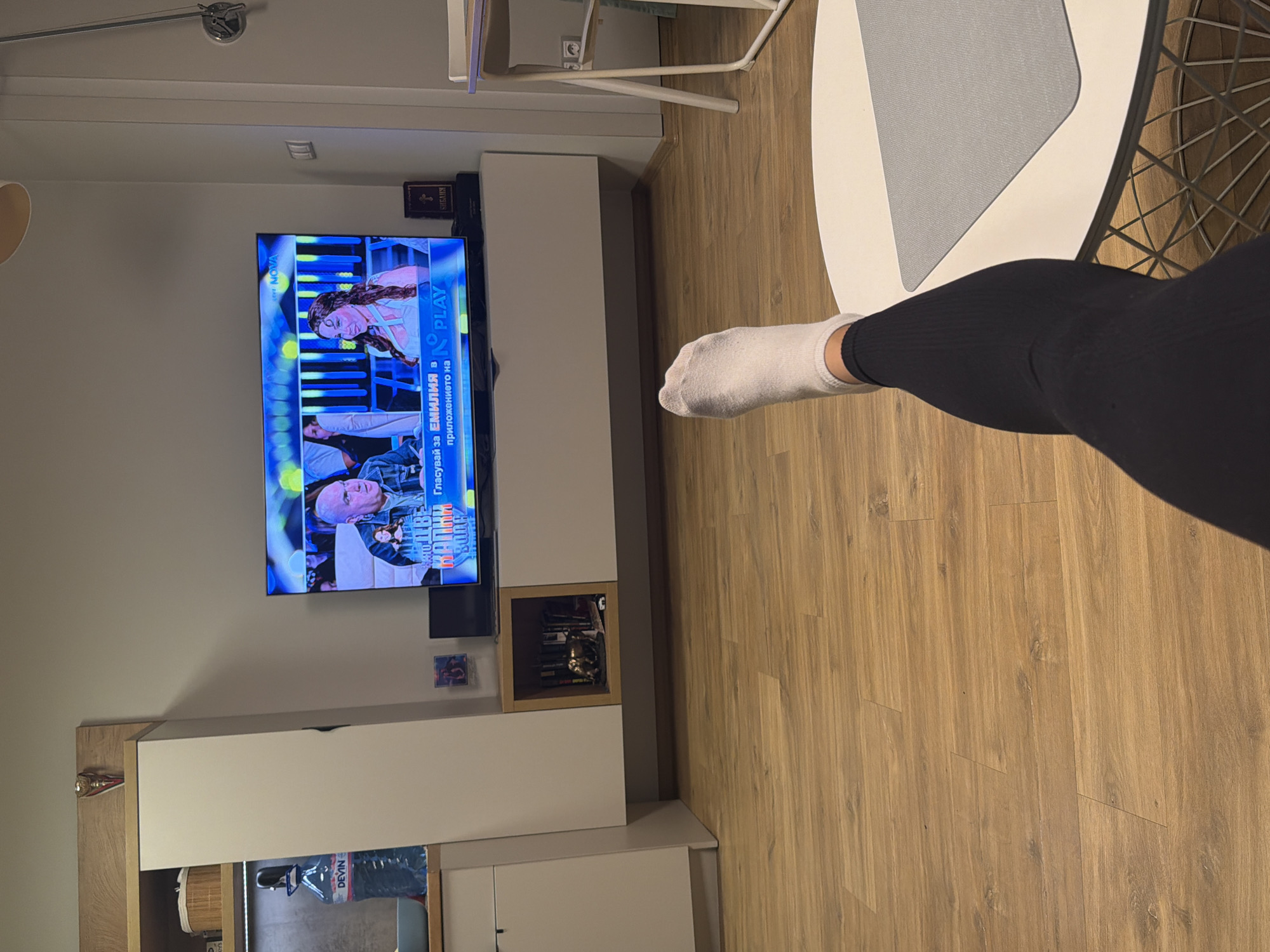 The image presents a first-person perspective of a relaxed indoor setting, likely a living room in Sofia, Bulgaria. In the immediate foreground, a person's left leg, clad in black ribbed leggings and a white sock, extends into the frame, suggesting a comfortable, possibly reclined, posture.
The primary focus of the scene is a wall-mounted television displaying a broadcast from "NOVA," a Bulgarian television channel. The screen shows two individuals, a woman with long dark hair and an older bald man, indicative of a show or competition. Prominently displayed text on the screen reads: "ГЛАСУВАЙ ЗА ЕМИЛИЯ В NOVA PLAY приложението на..." (Vote for Emiliya in NOVA PLAY app...).
Below the television is a modern media console, featuring white and light wooden elements. To the right of the TV, a small dark object, possibly a decorative book, is visible, bearing the Cyrillic text "БИБЛИЯ" (Bible) on its spine. Further down the console, an open shelf contains several books and a small golden statuette. Another visible item is a bottle of "DEVIN" brand water on a lower shelf.
The room features light brown wooden laminate flooring. Partially visible in the upper right corner are segments of a white and grey table with a distinctive geometric wire-frame chair. A wall-mounted light fixture is visible near the top left. The ambient lighting suggests it is likely evening or nighttime, creating a cozy and casual atmosphere.