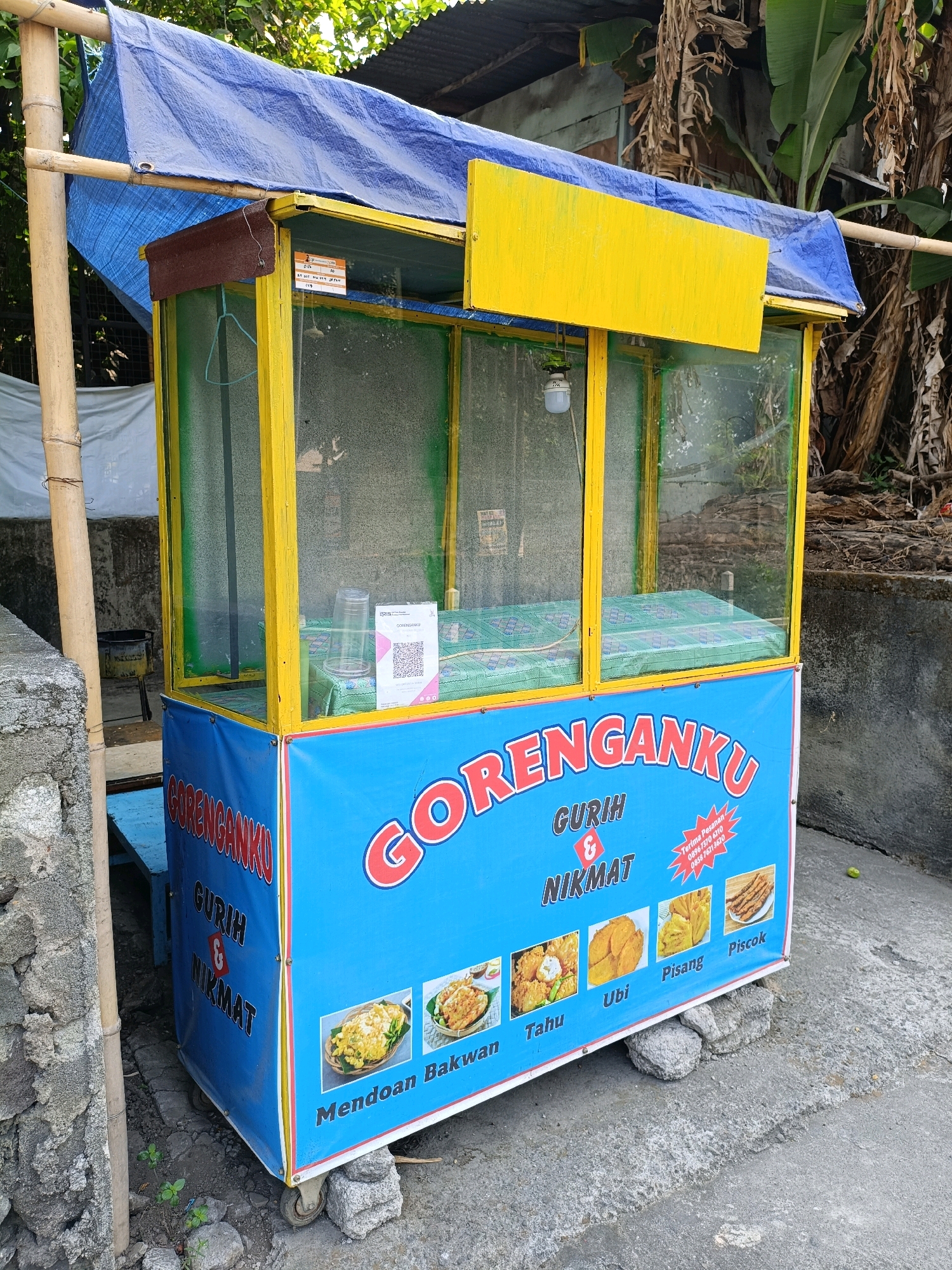 This is a full-shot, eye-level, outdoor photograph of a food cart in Maguwoharjo, Indonesia. The cart is blue and yellow, with a blue tarp covering the top. The cart has glass windows on the front and sides, revealing a green cloth inside. The front of the cart has the words "GORENGANKU" in large, red letters. Below this, in smaller text, are the words "GURIR & NIKMAT." There are also images of fried food items, including "Mendoan Bakwan," "Tahu," "Ubi," "Pisang," and "Piscok." A QR code is visible on the inside of the cart. The cart is on a concrete surface and is propped up on rocks. The background shows trees and a weathered wall. The scene is well-lit, suggesting it was taken during the daytime.