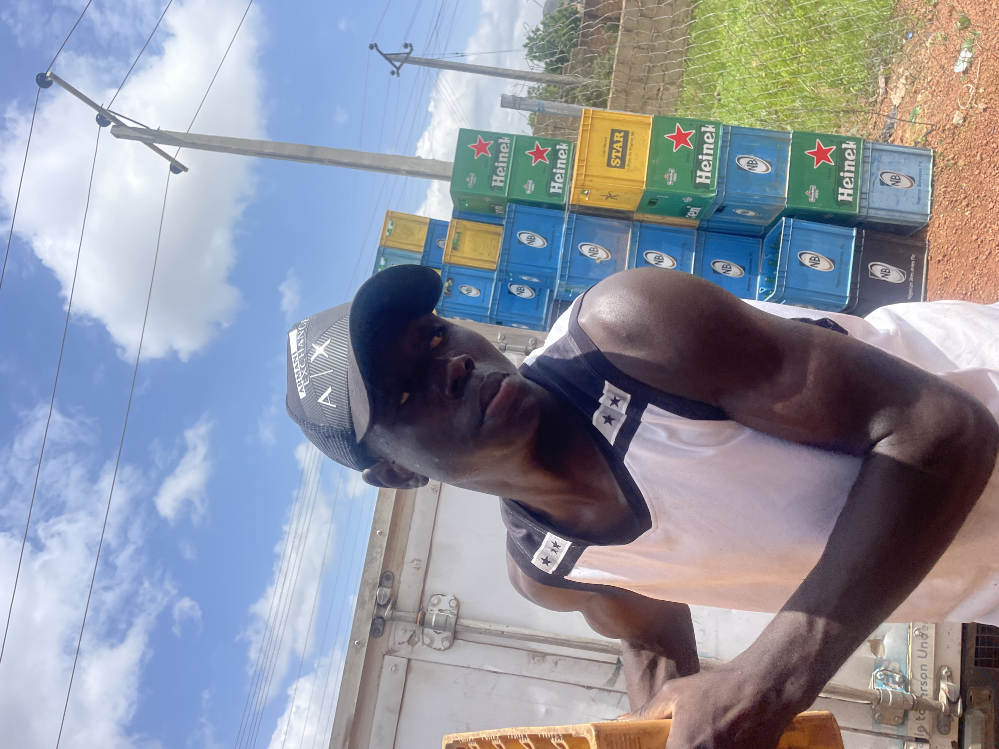 A young Black man wearing a black baseball cap and a white and black tank top stands outdoors in Gura Top, Nigeria, with stacks of crates behind him. The man is looking at the camera, his head slightly tilted to the side. His left arm is bent at the elbow and rests on a yellow crate. His right arm is down by his side. Behind the man are stacks of various beer crates, including green Heineken crates, yellow Star beer crates, and blue NB crates. The crates are stacked several high, forming a wall to his right. In the background, there is a utility pole with wires and a cloudy blue sky. The ground appears to be dirt with some grass and a chain-link fence. A white truck is visible to the left of the man. The scene suggests an outdoor storage or distribution area for beverages.